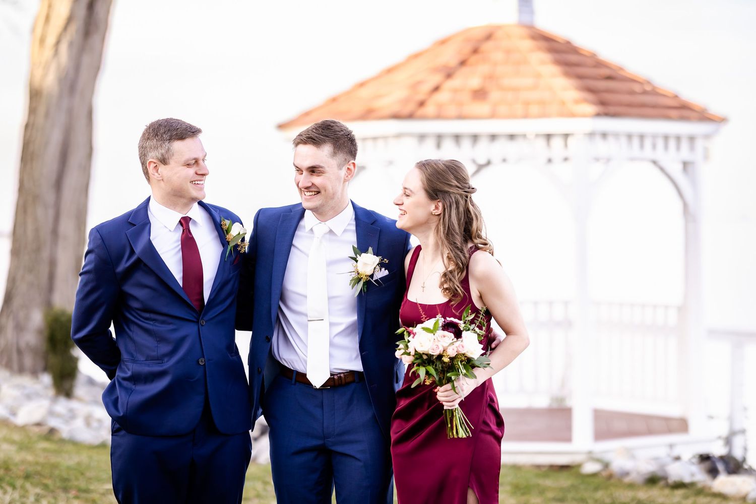 Wedding party in navy suits and burgundy dresses poses outdoors for formal photos.