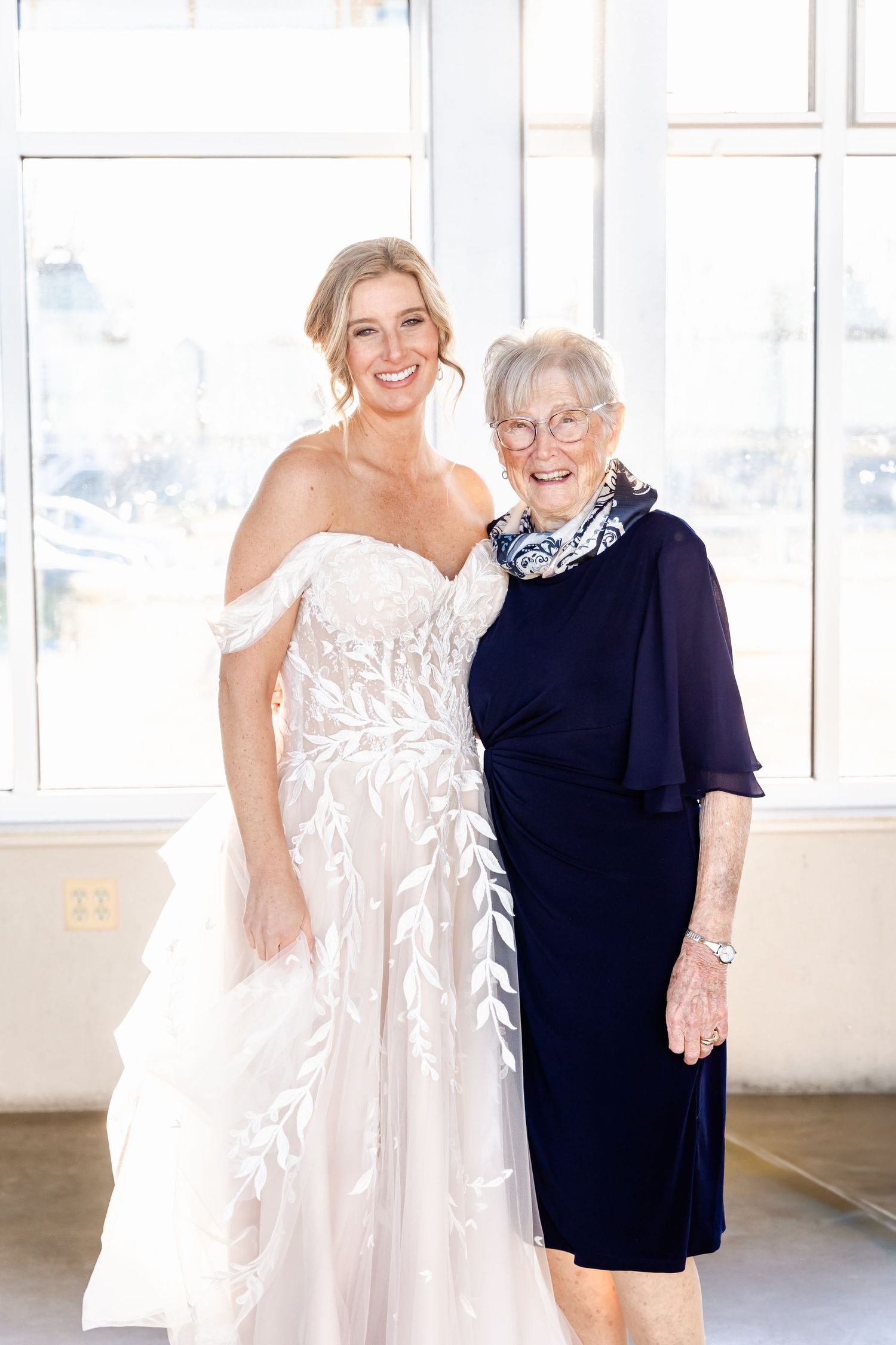 Elegant bride in off-shoulder lace gown poses with companion wearing navy dress by bright windows.