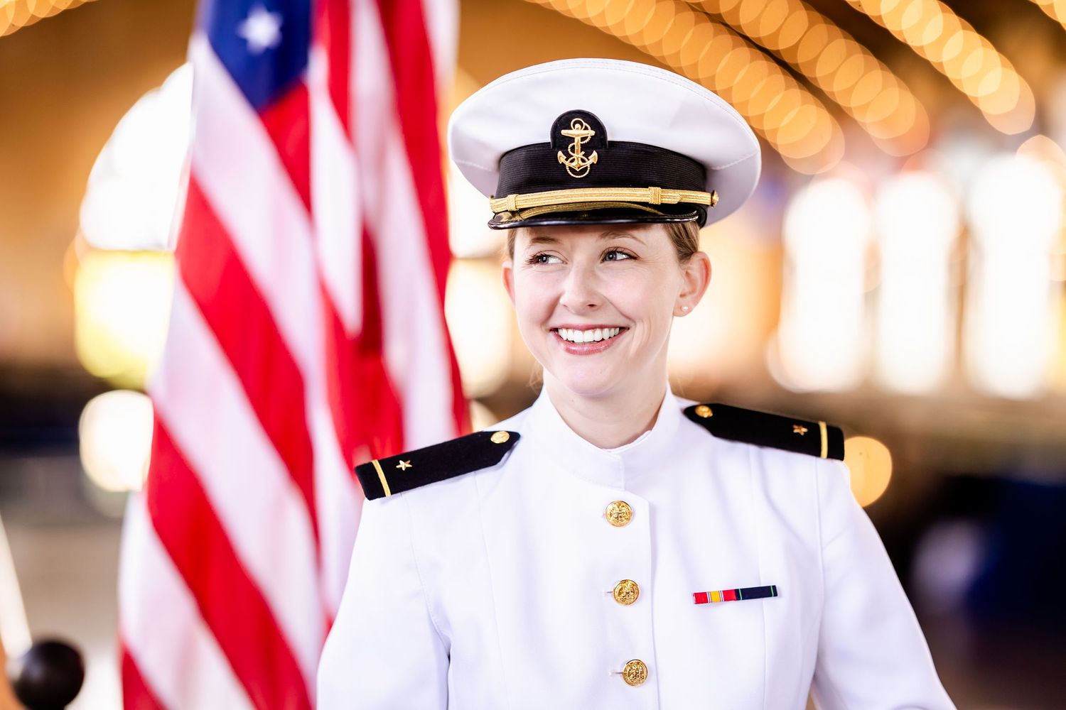 A service member in white naval dress uniform stands smiling with an American flag in the background.