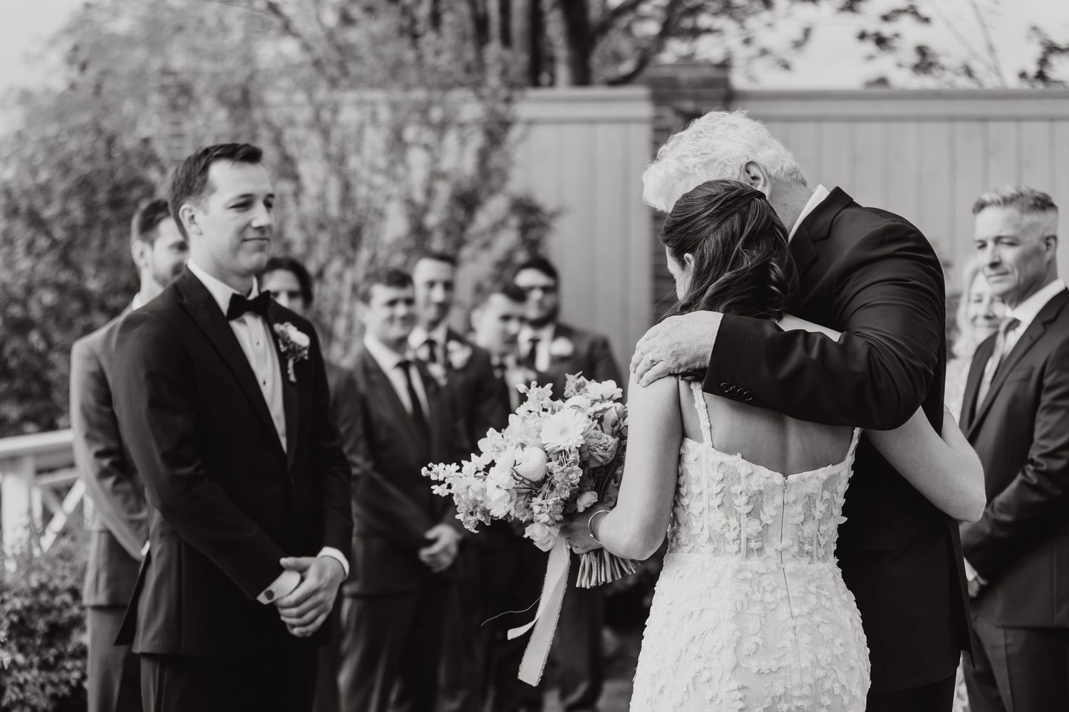 Black and white photo of an outdoor wedding ceremony showing couple sharing first kiss while officiant looks on with smile.