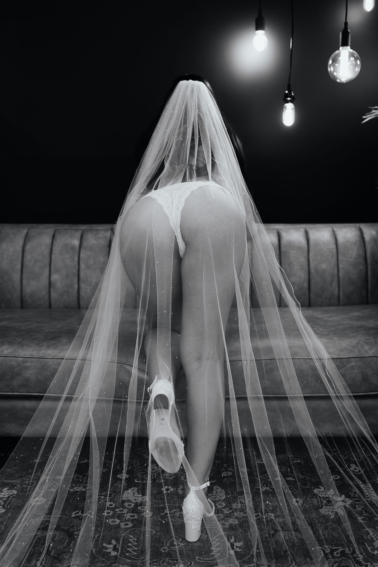 Black and white photo of a bride in white lingerie and veil kneeling on a sofa with decorative lights above.
