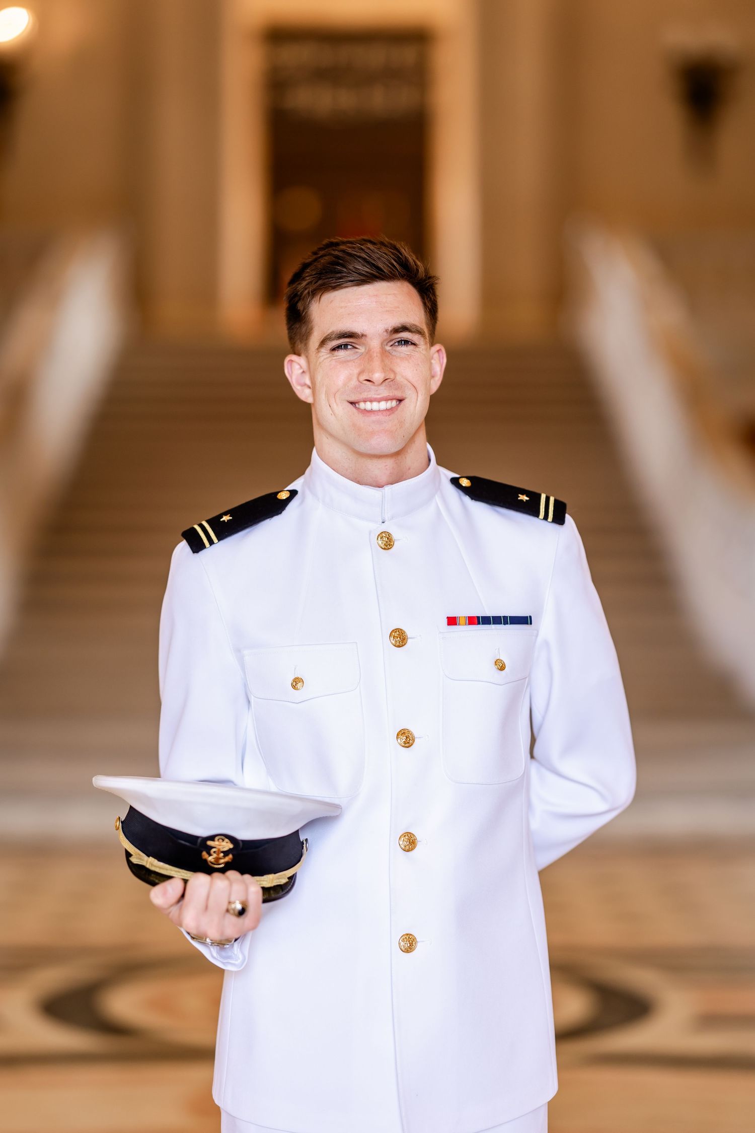Young naval officer in white dress uniform holds cap while smiling in elegant hall.