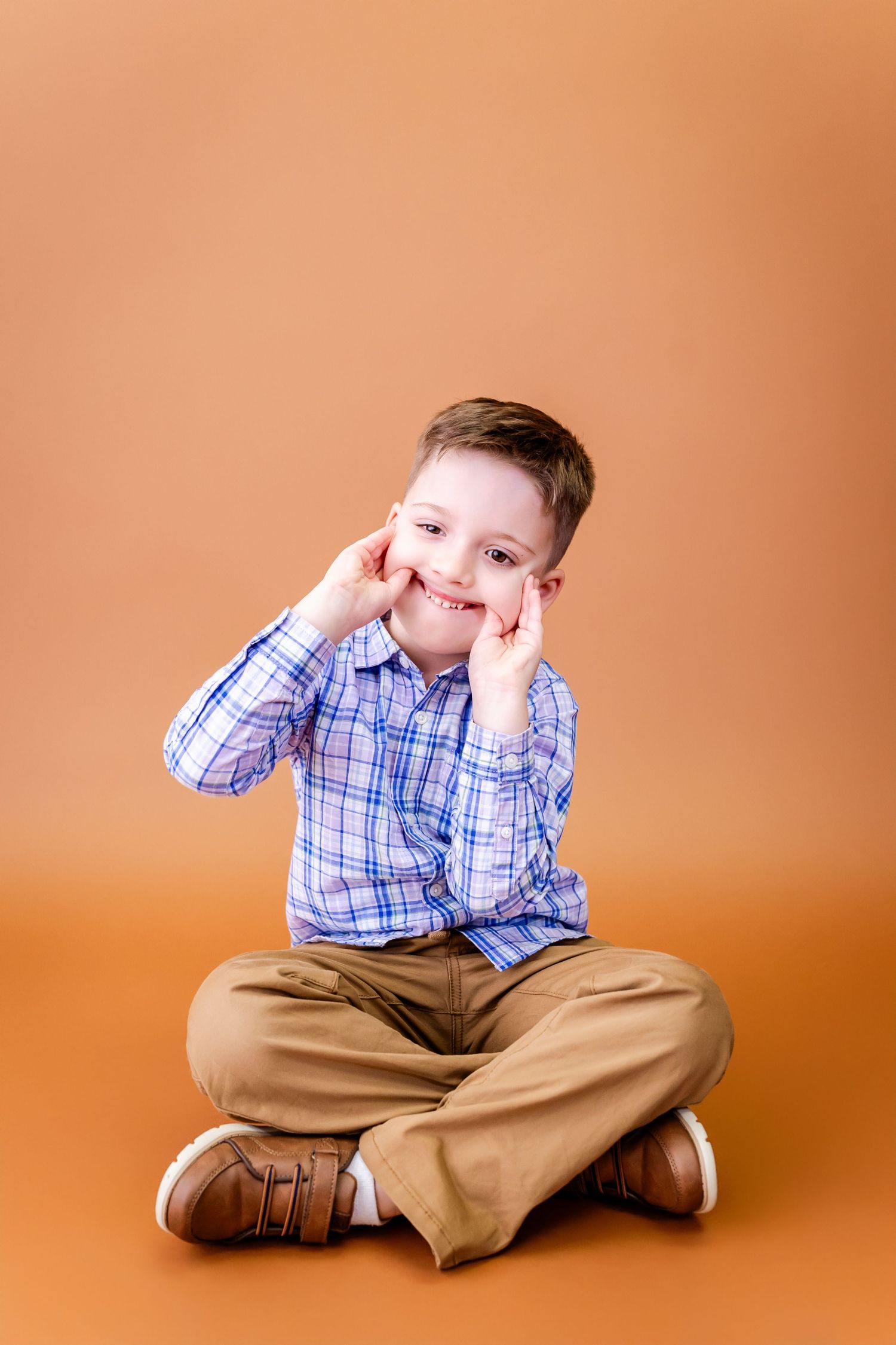 A child in a plaid shirt and khakis sits cross-legged against an orange background making silly expressions.