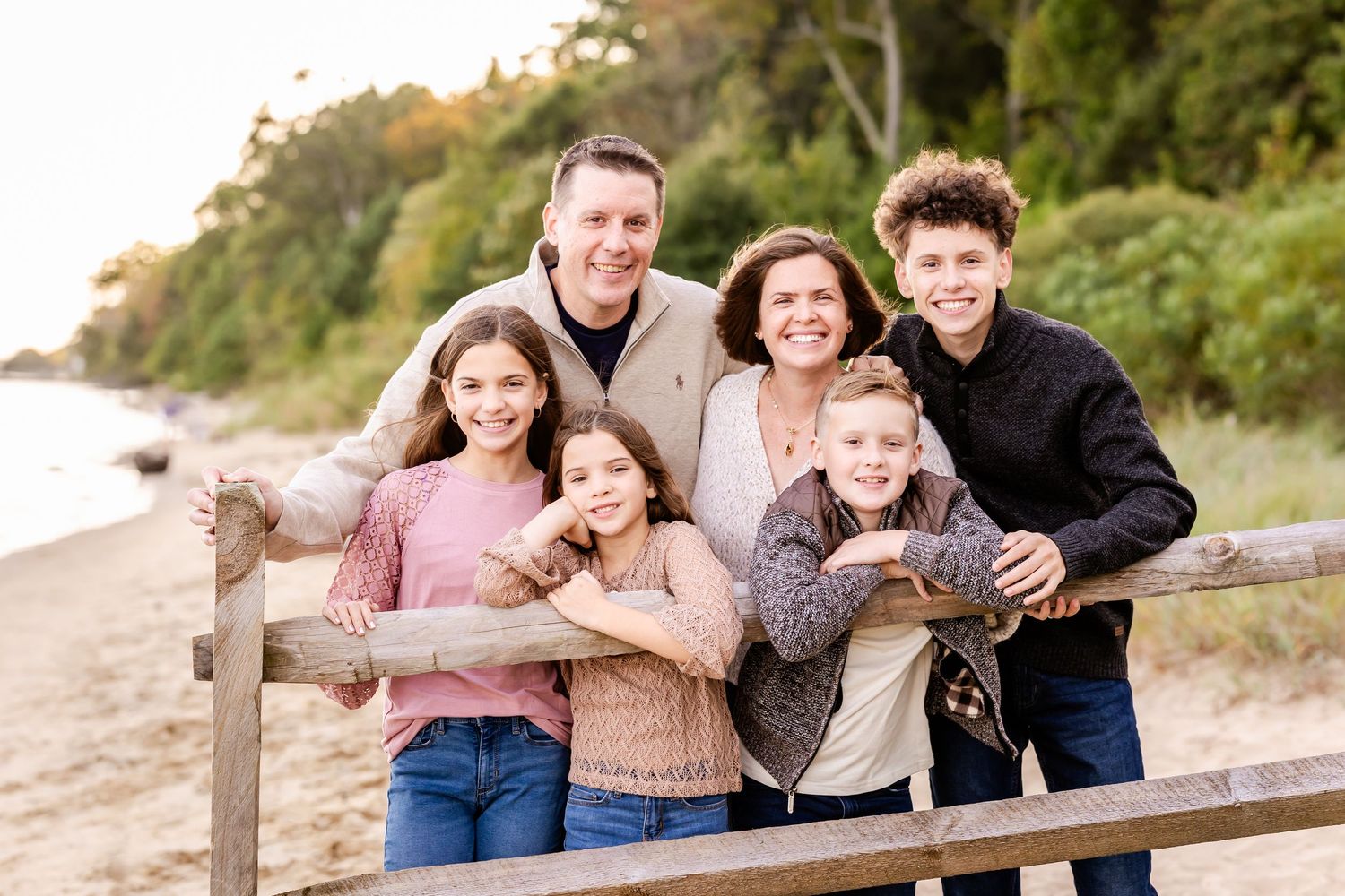 A happy family poses together on a wooden railing at the beach during golden hour with trees in the background.