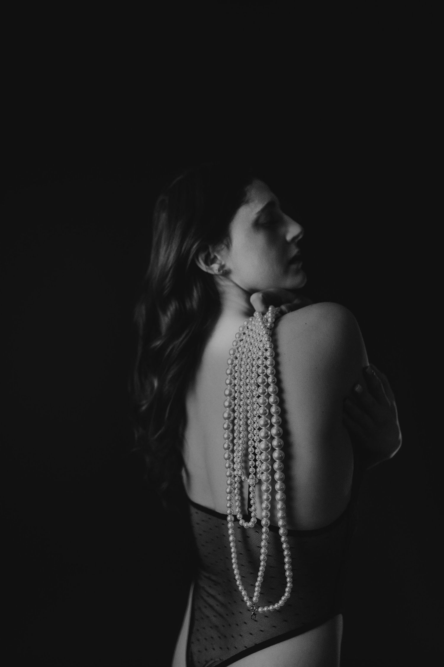Dramatic black and white portrait of a woman in dark attire with pearl strands draped down her bare back.