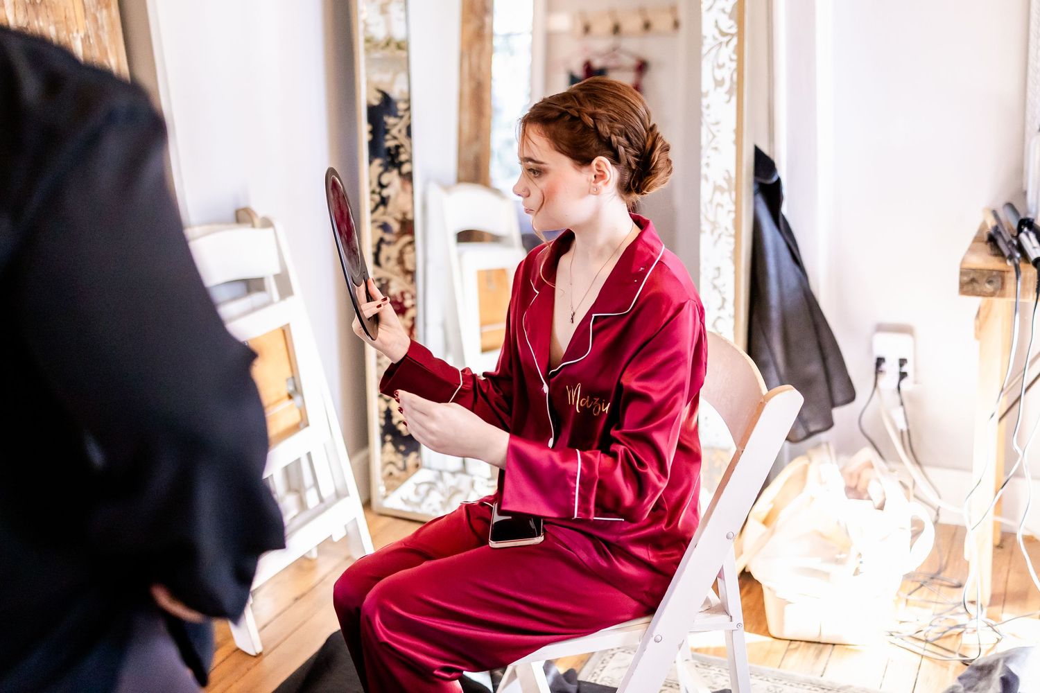 A person in a luxurious red silk pajama set sits while holding a mirror in a bright, elegant dressing room.