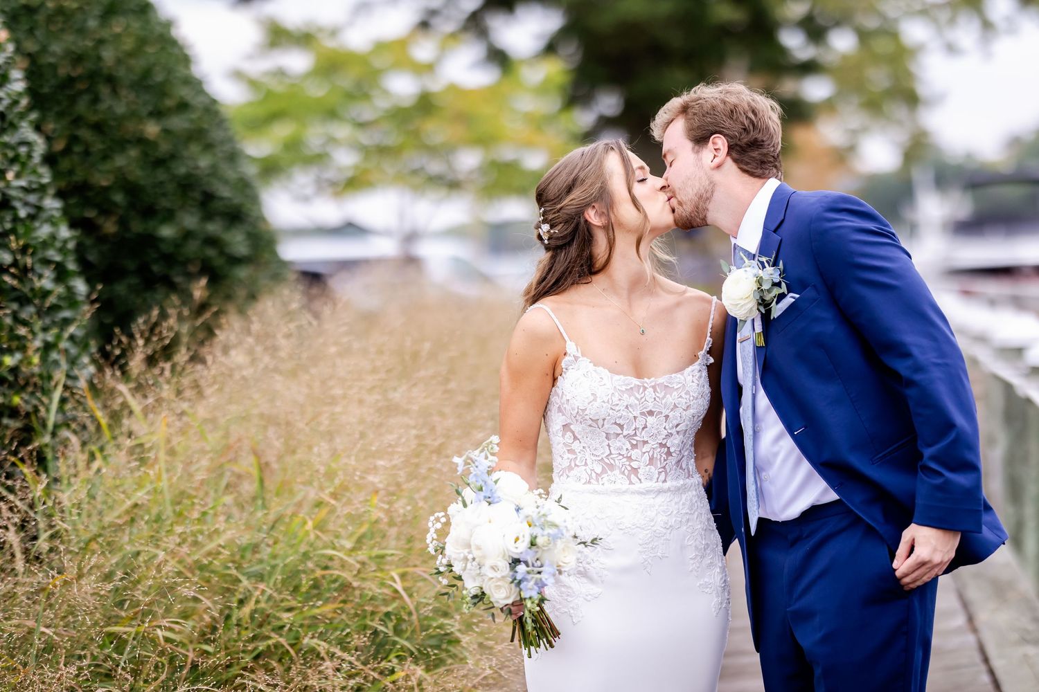 Wedding couple poses together on grassy path near waterfront in elegant attire.