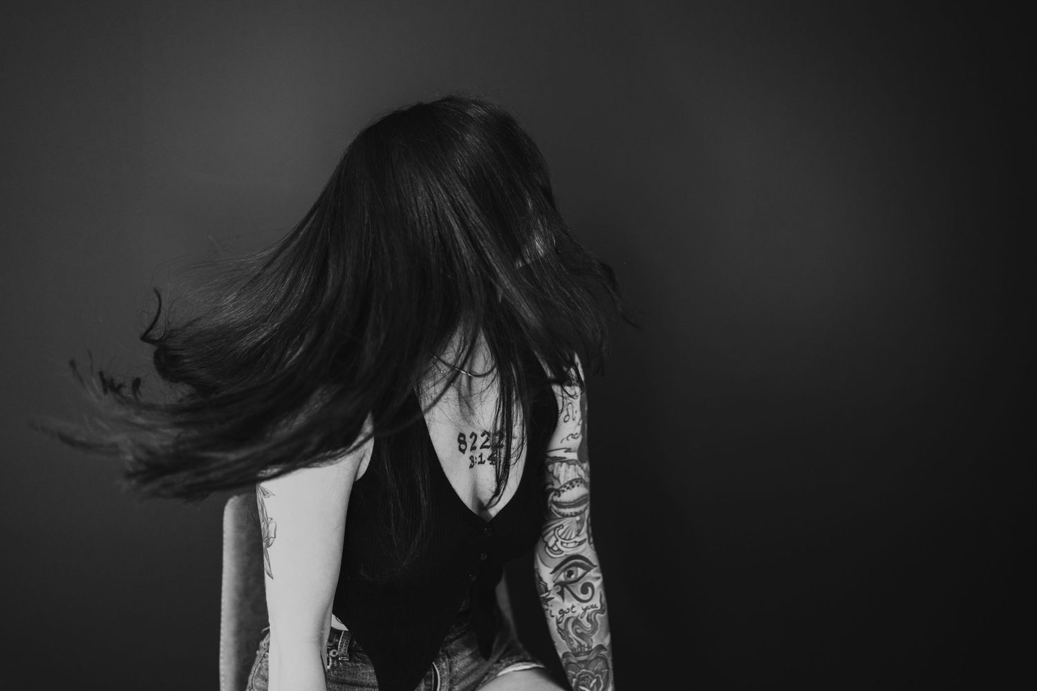 Black and white photo of a woman with long dark hair blowing, showcasing intricate tattoos on her arm and side.