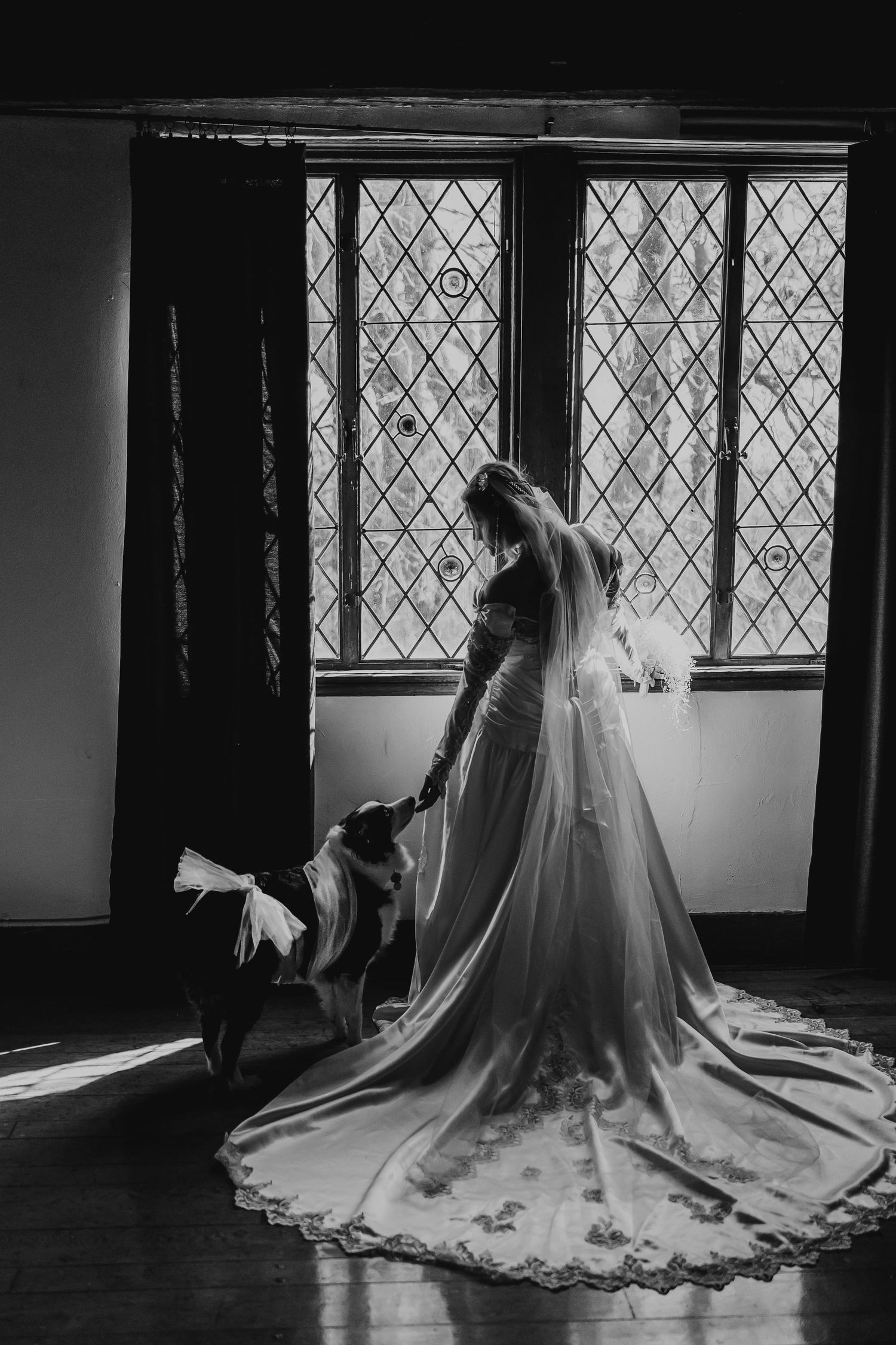 Bride in elegant wedding gown with long train stands by leaded glass windows in black and white.