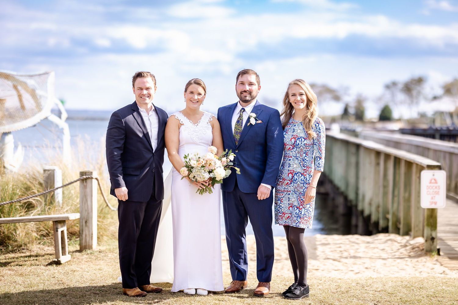 Bride and groom pose with two guests near waterfront dock on sunny wedding day.