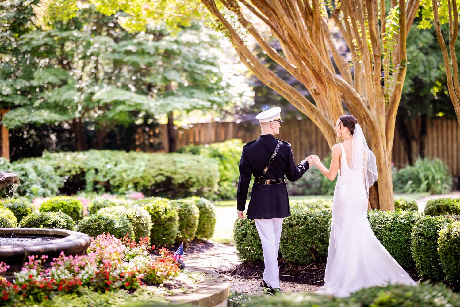 A couple stands together in a beautiful garden setting with manicured hedges and blooming flowers.