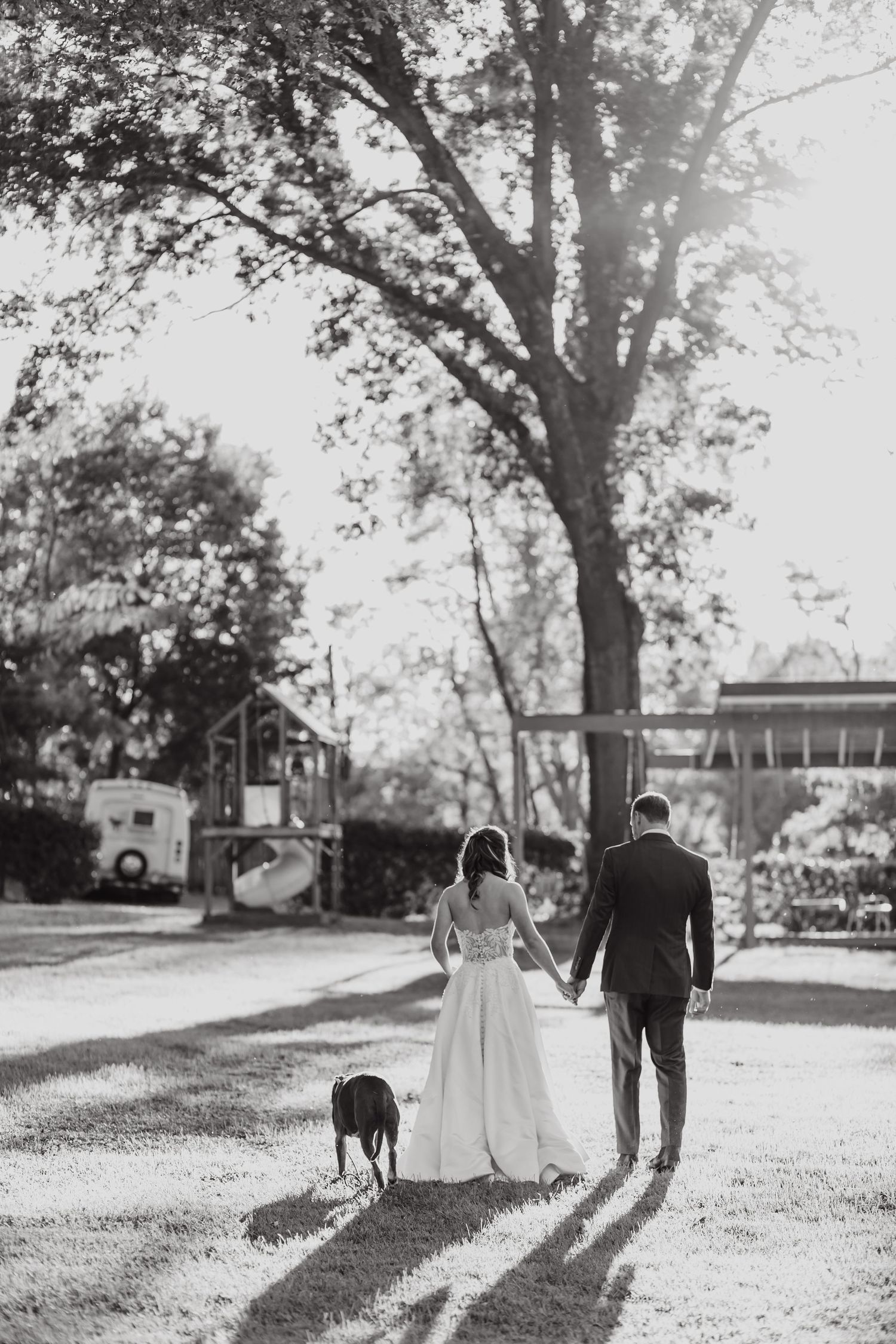 A black and white photo of newlyweds walking away together on a tree-lined path casting long shadows.