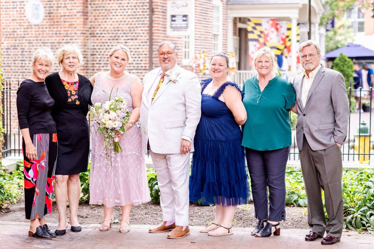 Wedding party members posing outdoors against brick building with decorative landscaping.