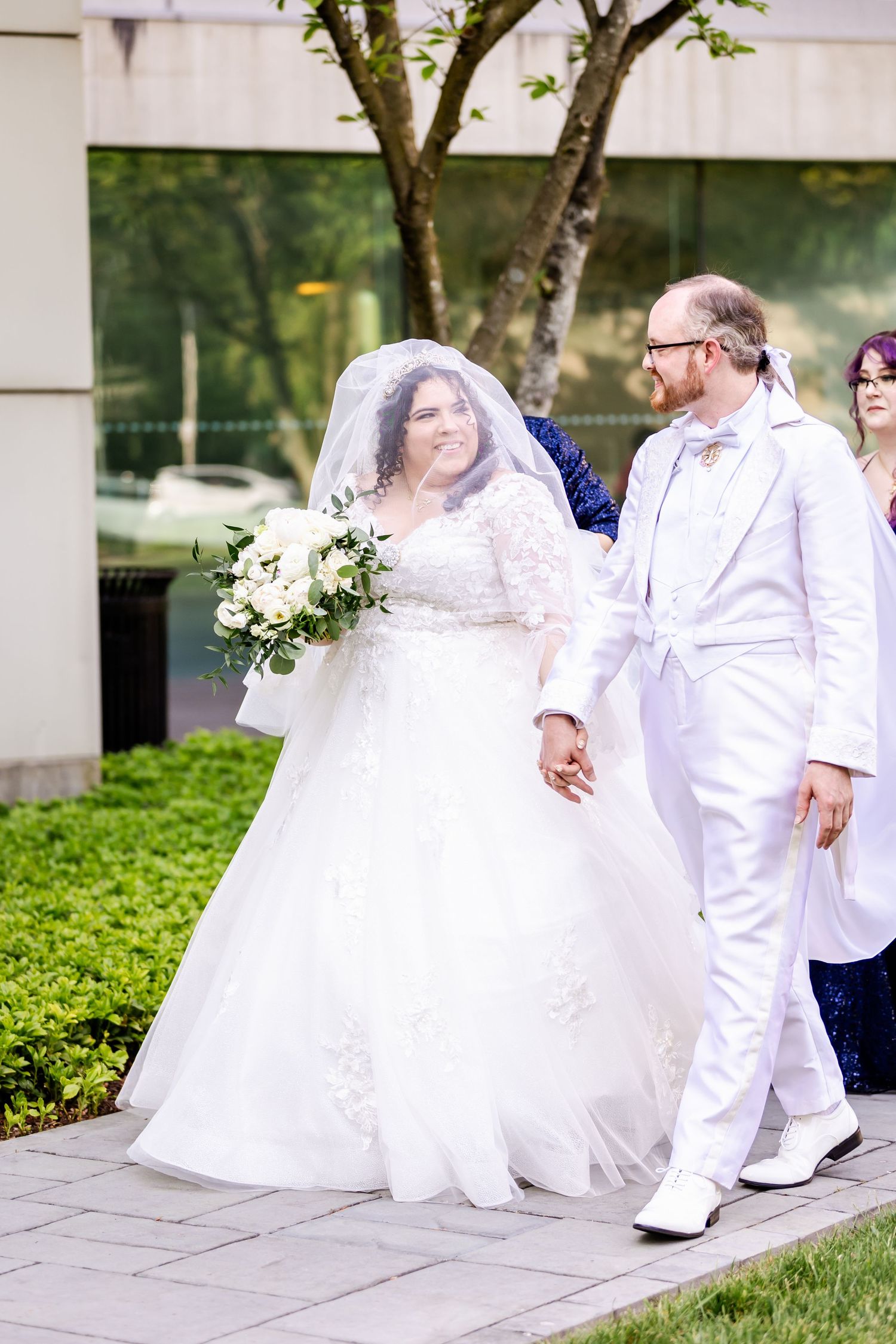 Wedding couple in white attire walk together holding hands outdoors.