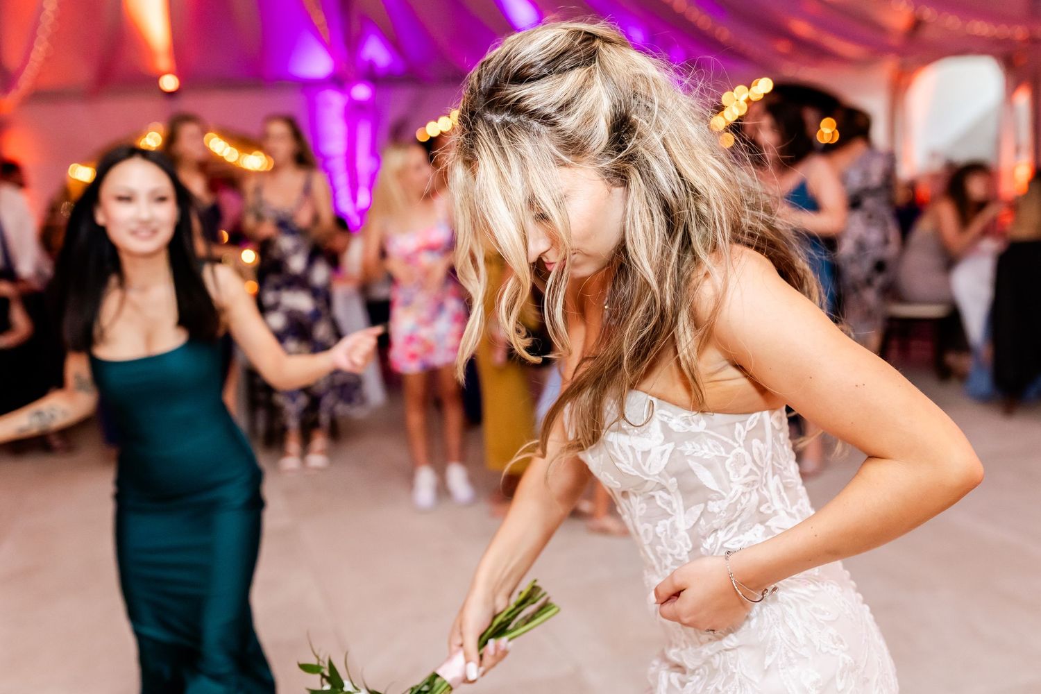 Person in a white lace wedding dress dancing energetically at a reception with purple lighting and guests in background.
