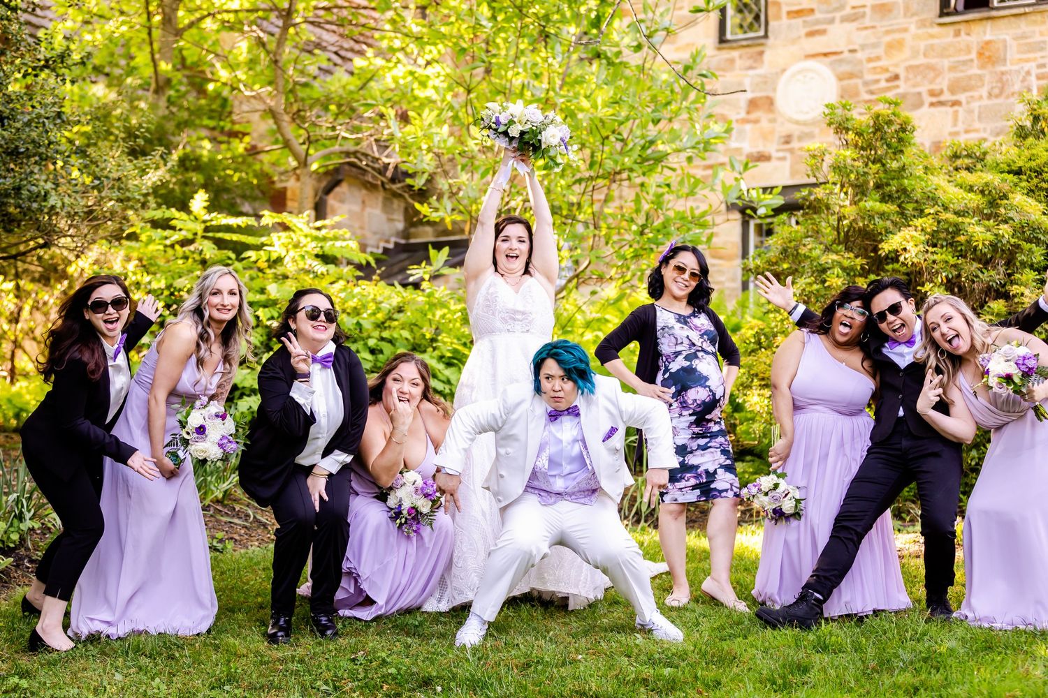 Wedding party in purple dresses and dark suits posing playfully with bouquets in a garden setting.