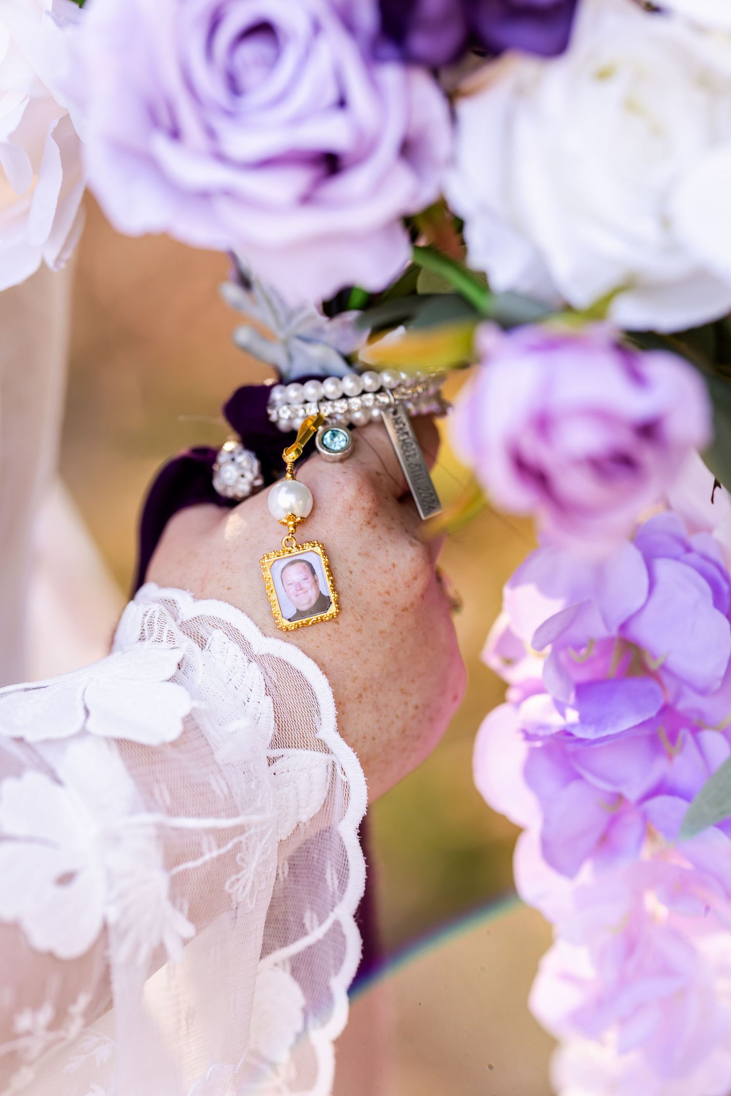 Close-up of a charm bracelet with locket and pearls surrounded by purple and white roses.