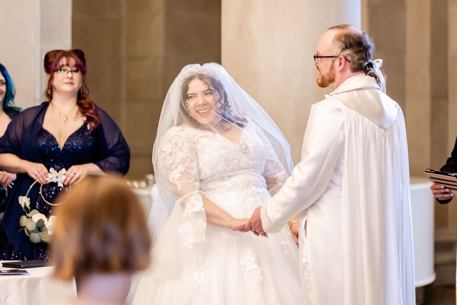 Wedding ceremony with bride in lace gown and officiant in white robes exchanging vows.