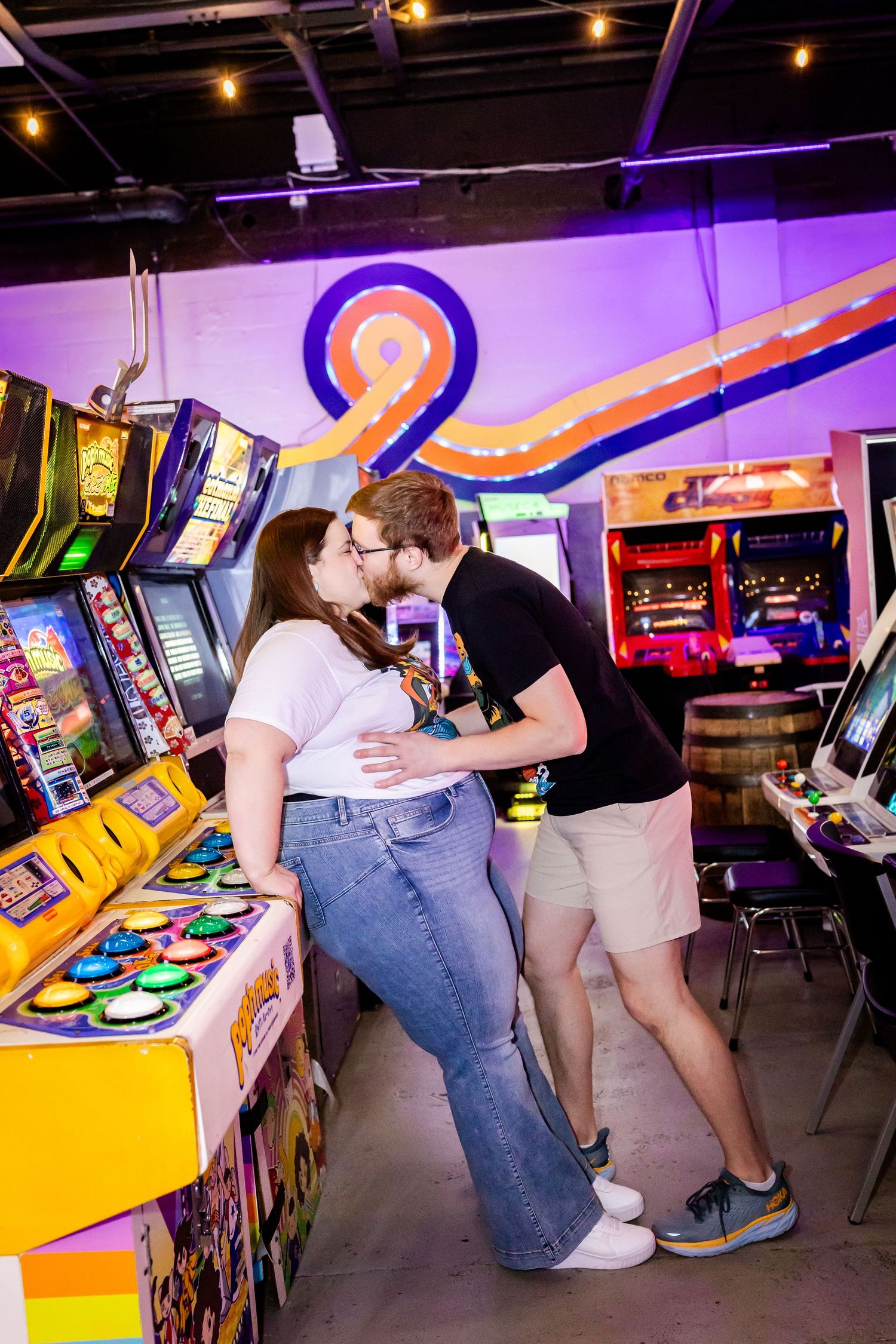 A couple shares a romantic moment while playing arcade games in a retro gaming venue with colorful neon lighting and decor.