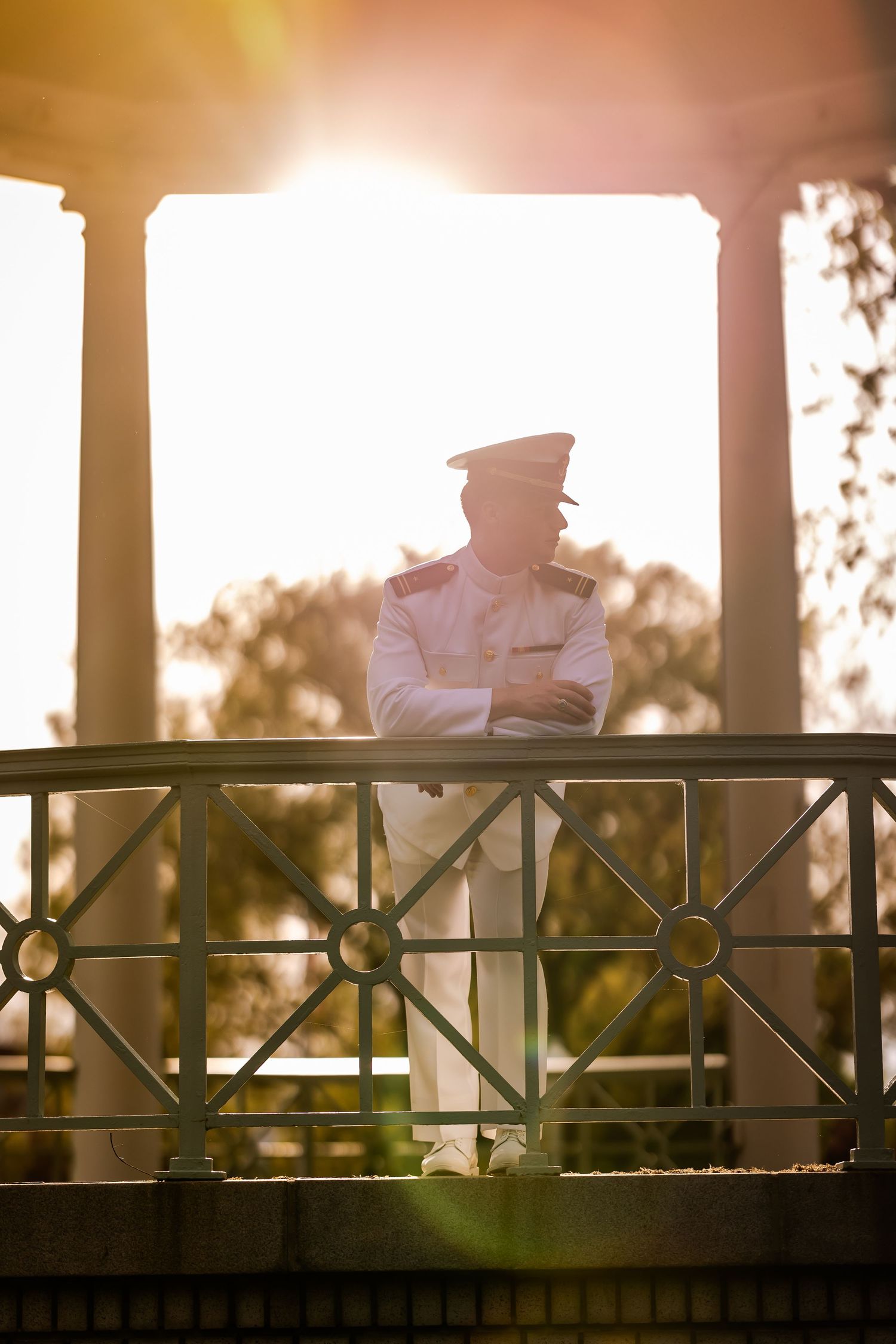 Naval officer in white uniform silhouetted against sunset while standing on ornate balcony.
