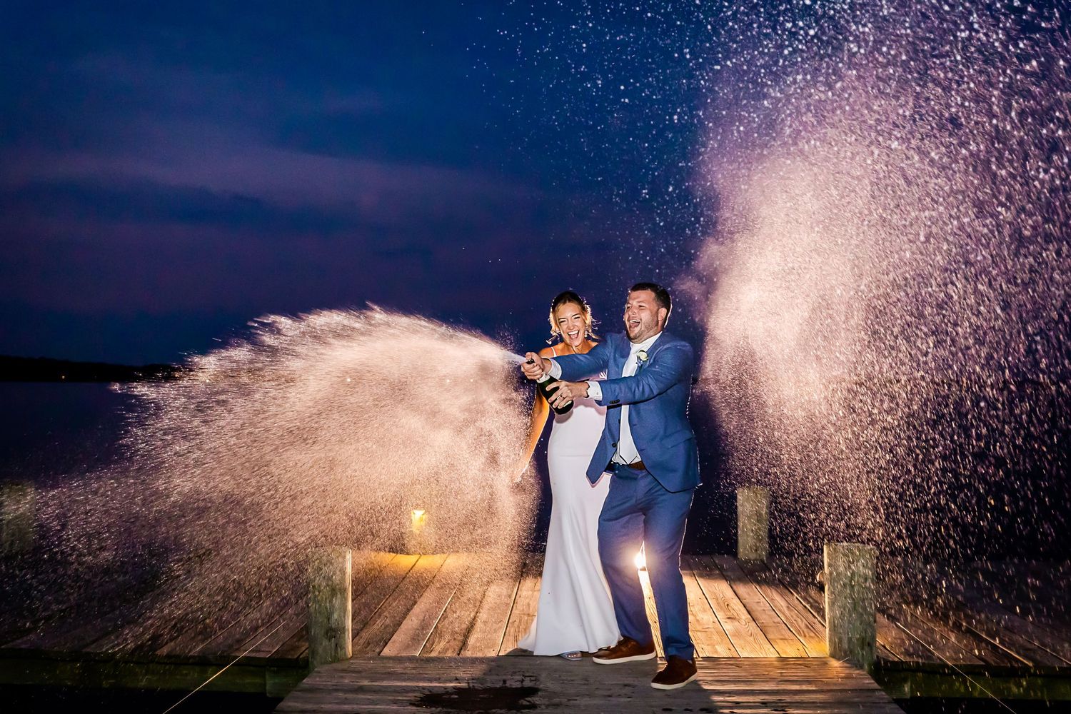A couple pops champagne on a wooden dock at night creating dramatic sprays against the dark sky and ocean.