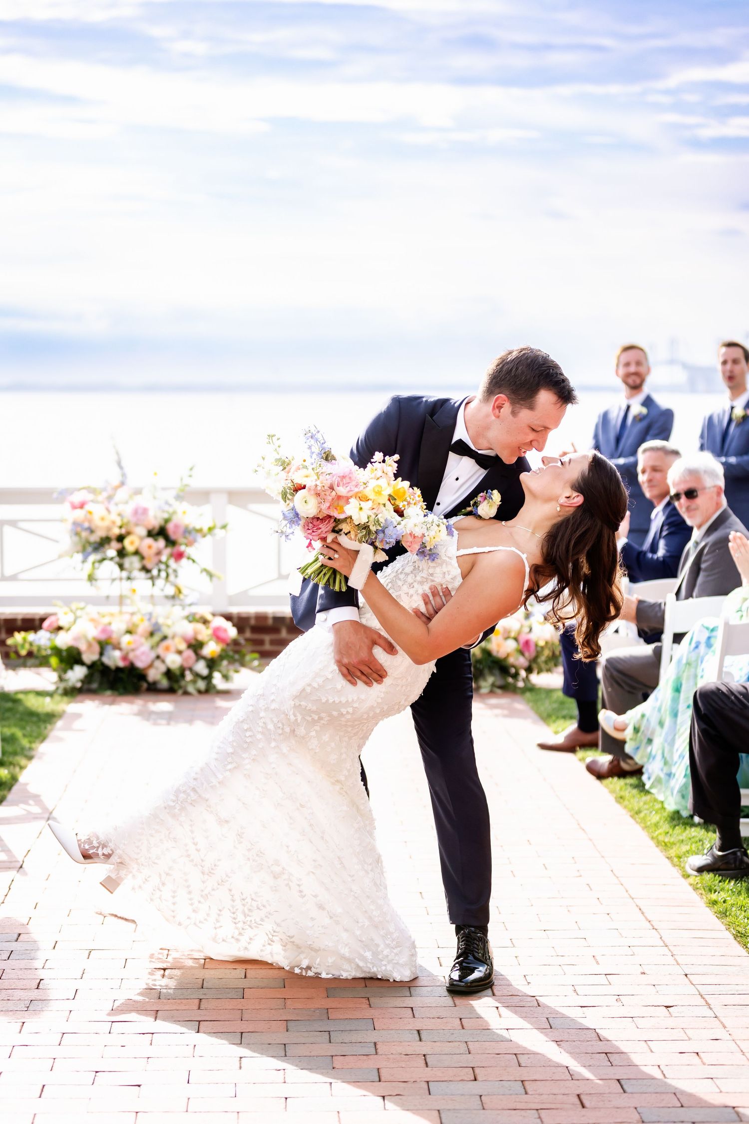 Romantic wedding couple shares a dip kiss on brick pathway with floral arrangements and guests in background.