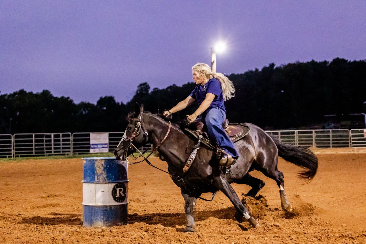 Rider competing in barrel racing on horseback at night in sandy arena with illuminated lights.