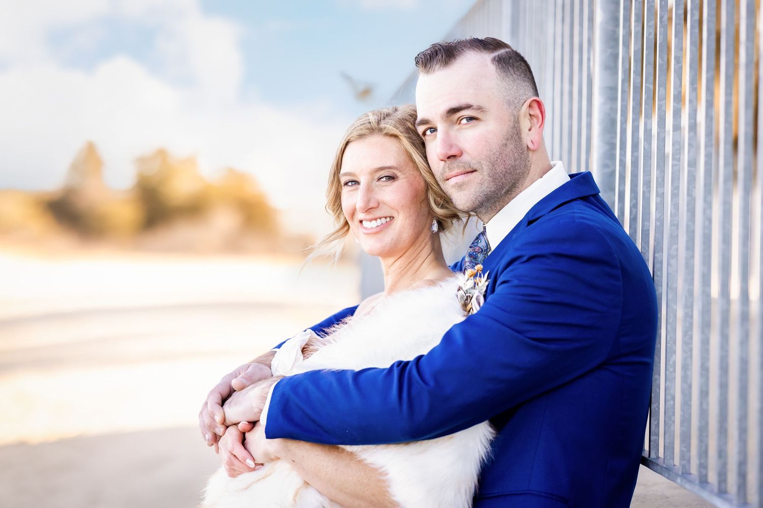 A couple in formal wedding attire poses together against an industrial backdrop.