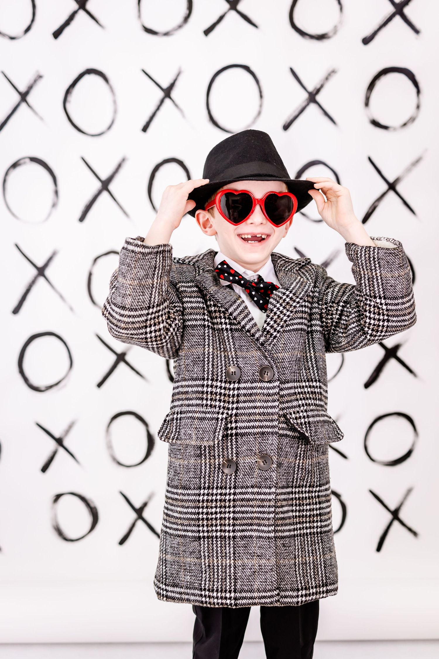 Stylish young person in plaid coat and heart-shaped sunglasses poses against XO pattern wall backdrop.