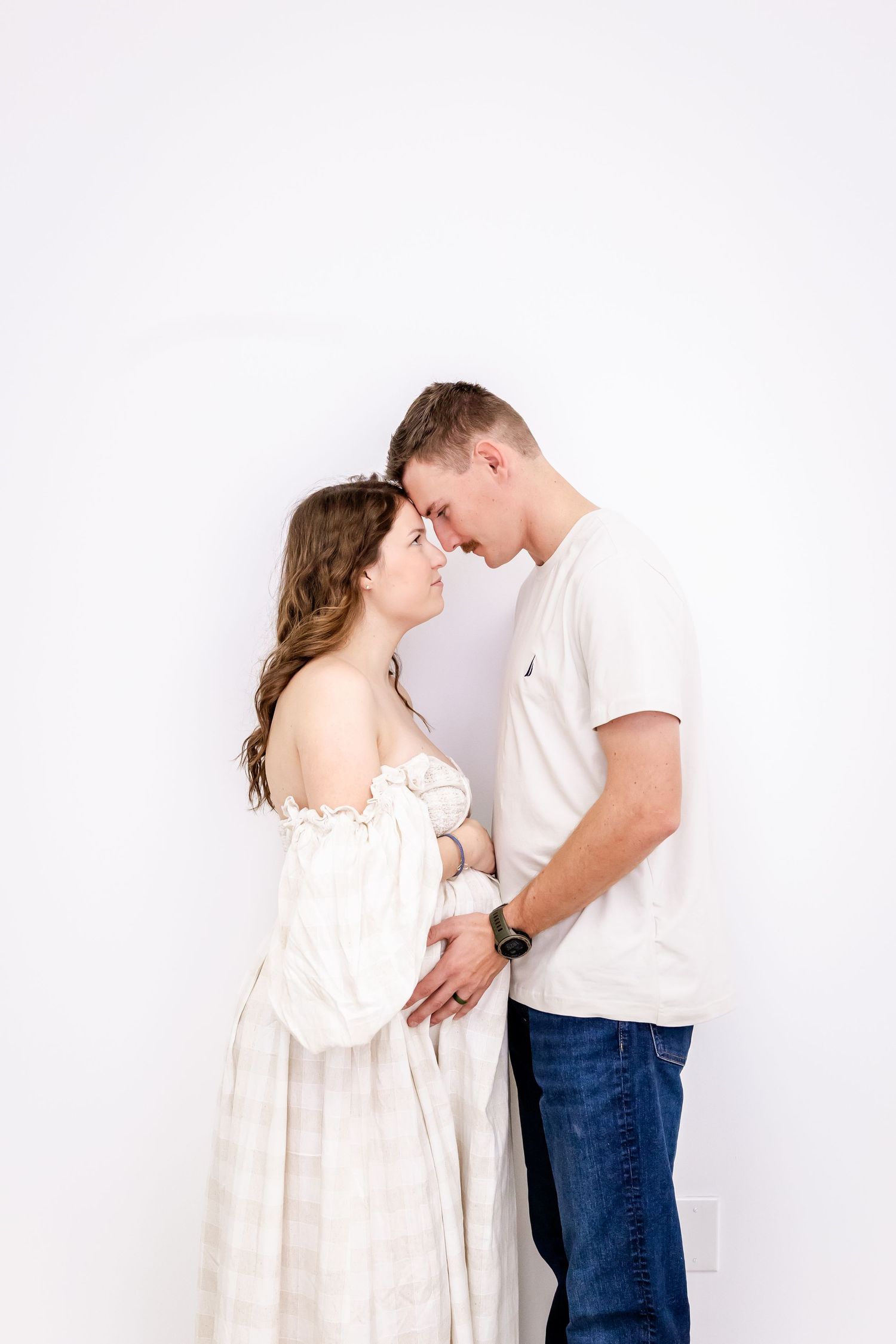 Intimate couple embraces in soft lighting against white backdrop with woman in flowy white dress and man in casual attire.
