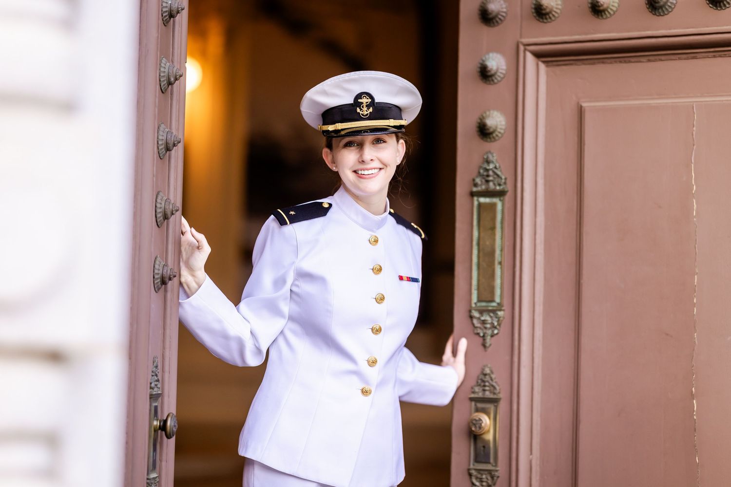 Naval officer in crisp white dress uniform and peaked cap stands smiling in decorative doorway.
