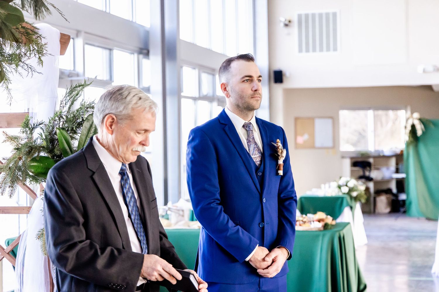 Two people in formal attire standing in bright indoor venue with green decor.