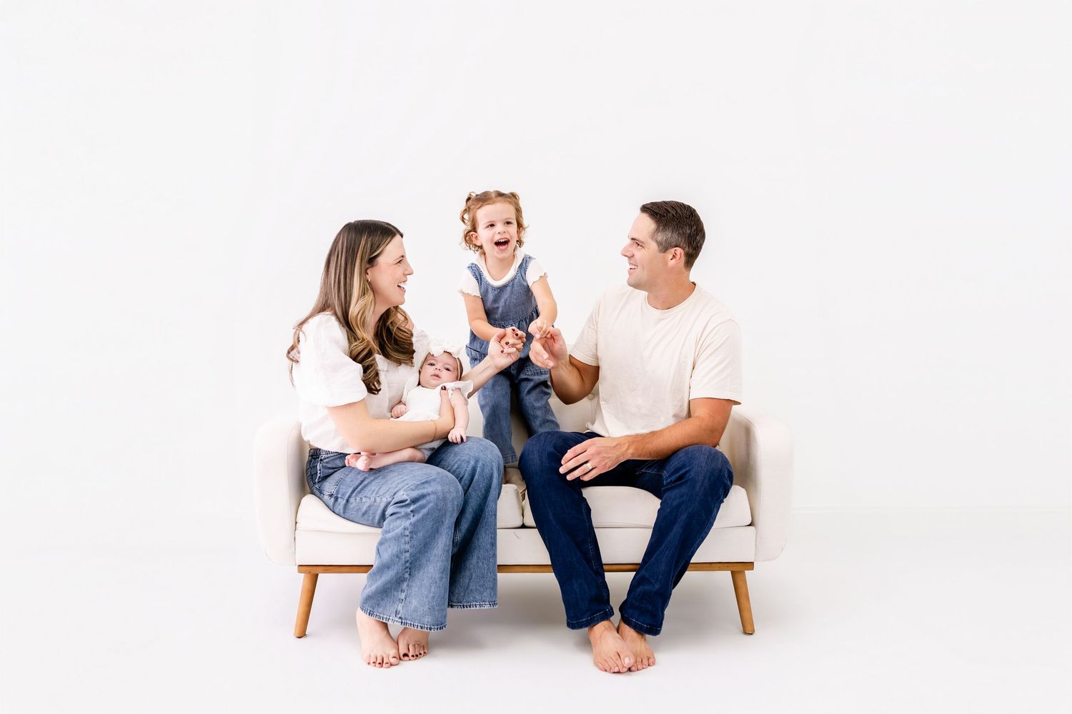 Smiling family of four sitting on a cream sofa against a white backdrop, with parents in jeans holding a newborn and a laughing toddler.