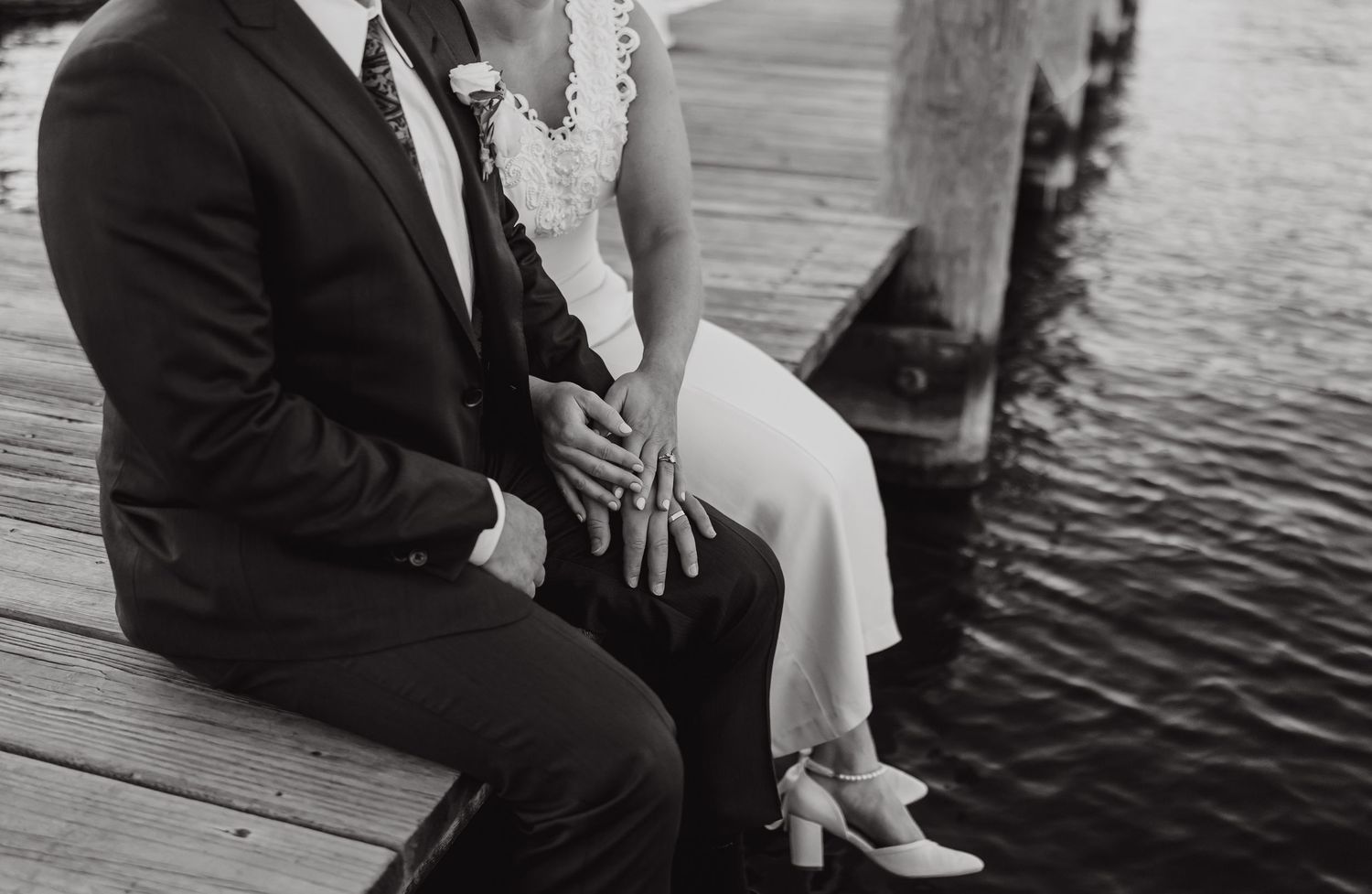 A romantic couple sits on a wooden dock by the water in formal attire during sunset.