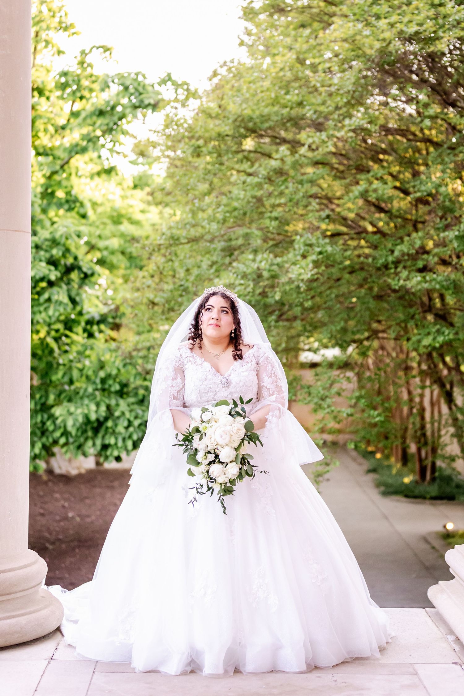 Bride in white gown with lace details poses with bouquet under outdoor garden archway.
