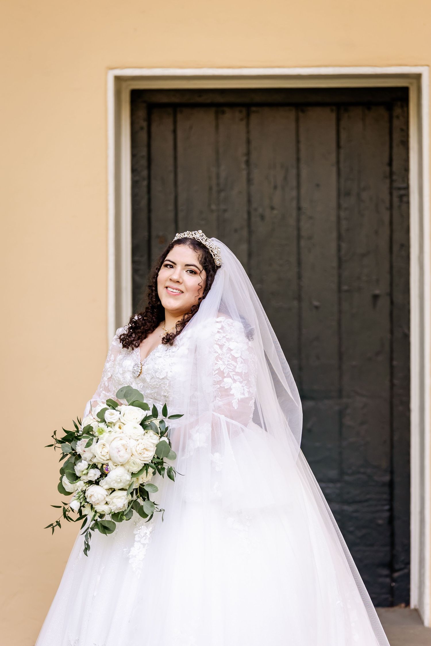 Bride in white lace gown holds elegant white floral bouquet against yellow colonial backdrop.