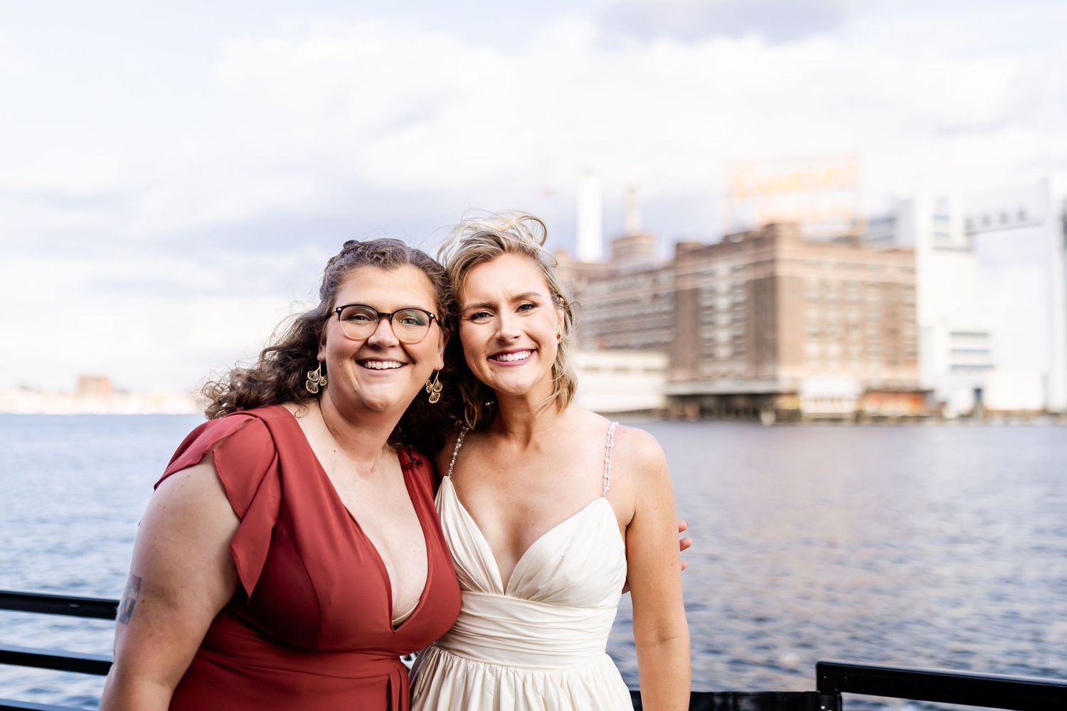 Two friends pose together on waterfront pier with harbor buildings in background.