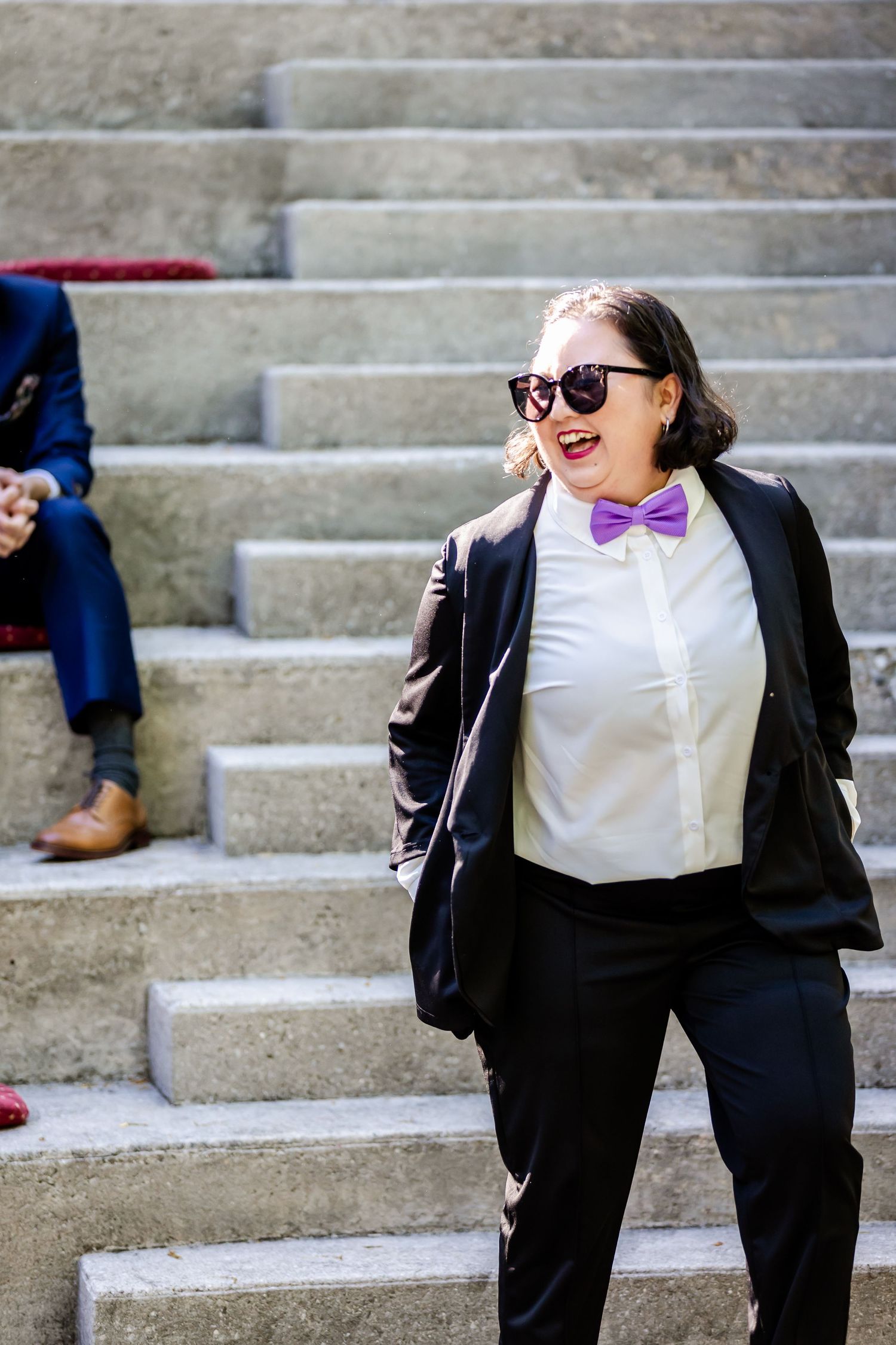 Person in black suit and purple bowtie smiles while standing on concrete steps.