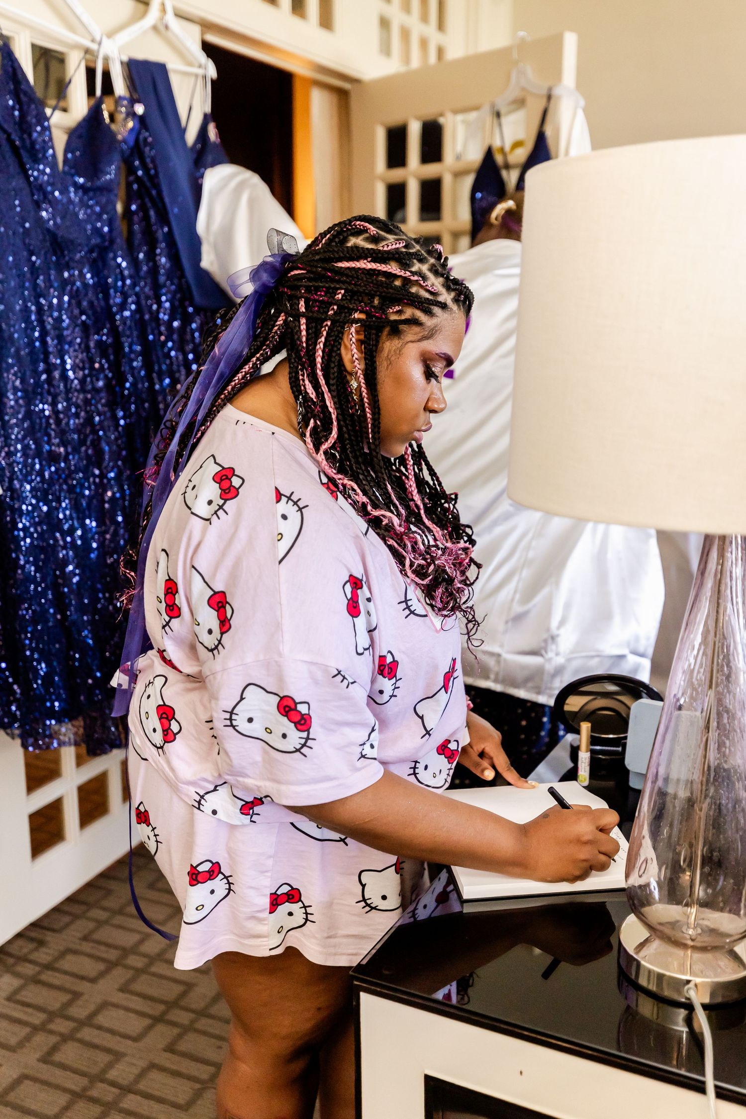 Someone writing in a Hello Kitty pajama shirt next to blue sequined dresses and a lamp.