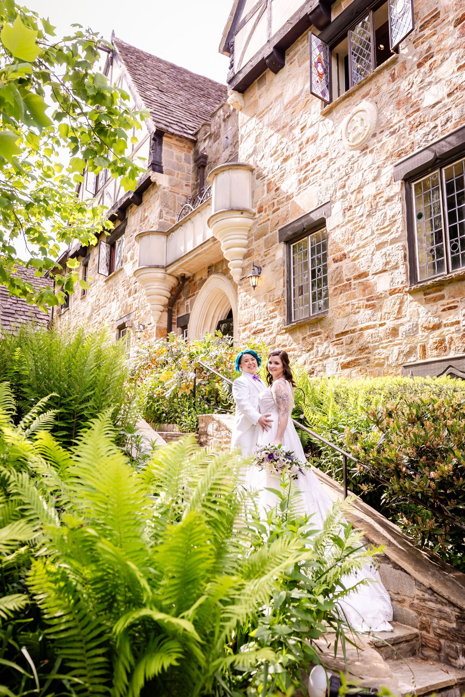 A wedding couple stands on stone steps outside a historic stone building with lush ferns and gardens.