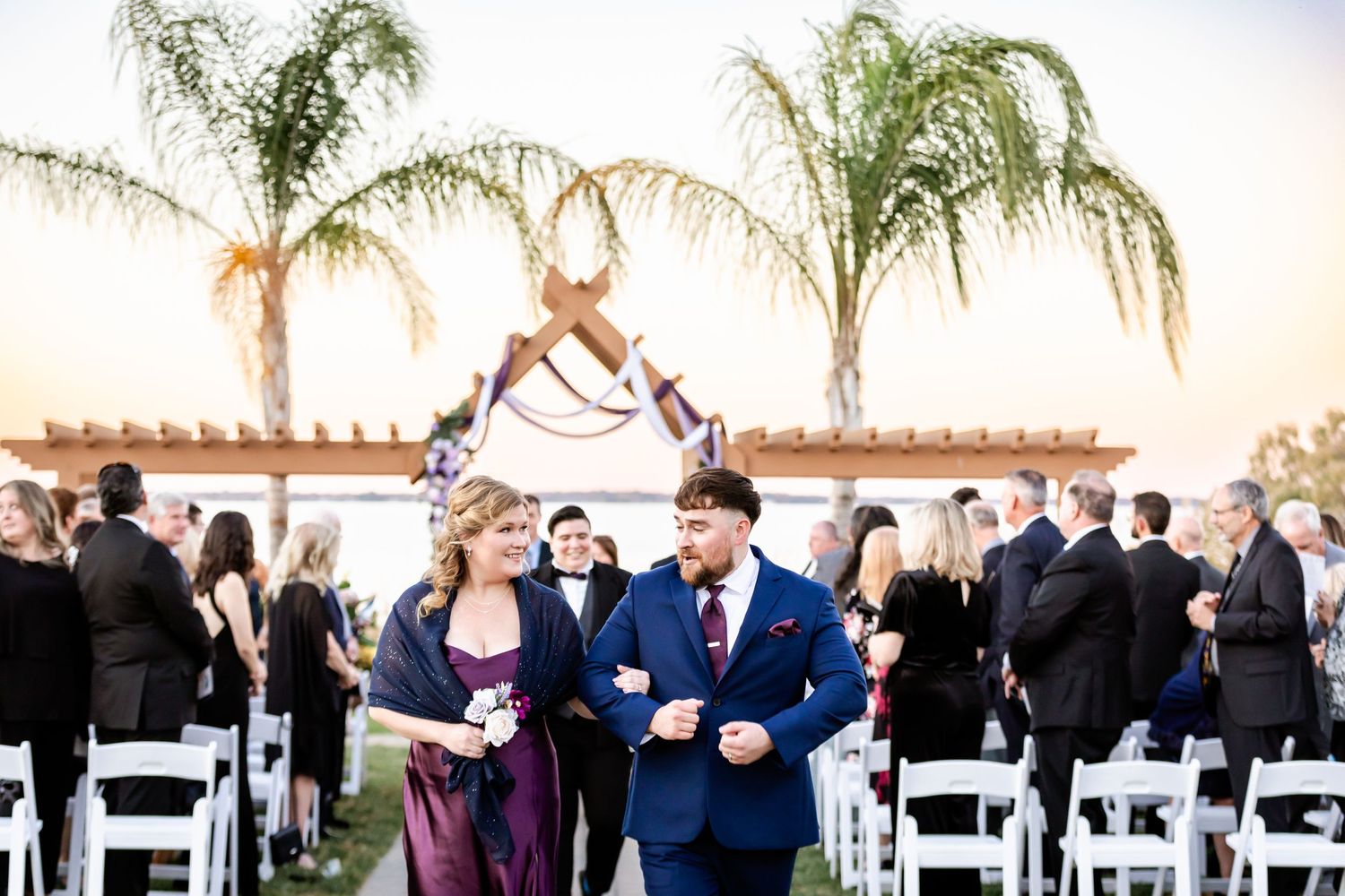 Wedding party walks between white chairs at outdoor ceremony with palm trees at dusk.