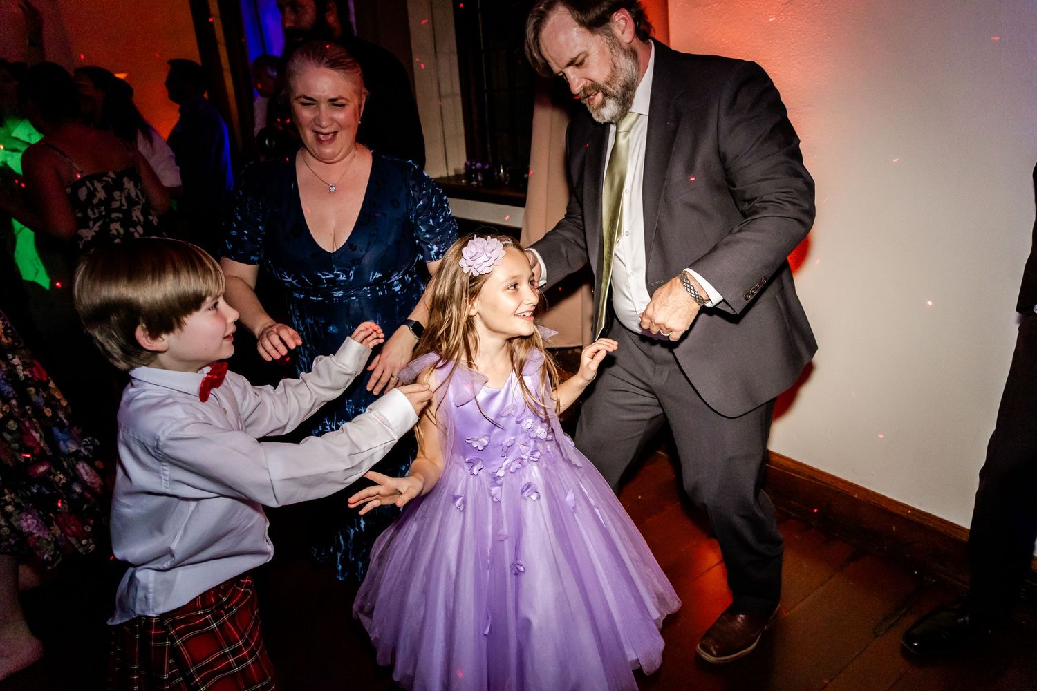 Young girl in purple dress dances with adults at wedding reception.