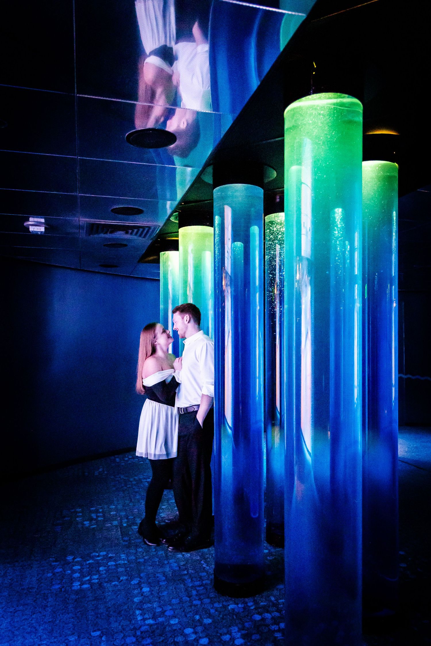 A couple embraces in a dark room with glowing blue and green illuminated cylindrical columns creating a moody atmosphere.