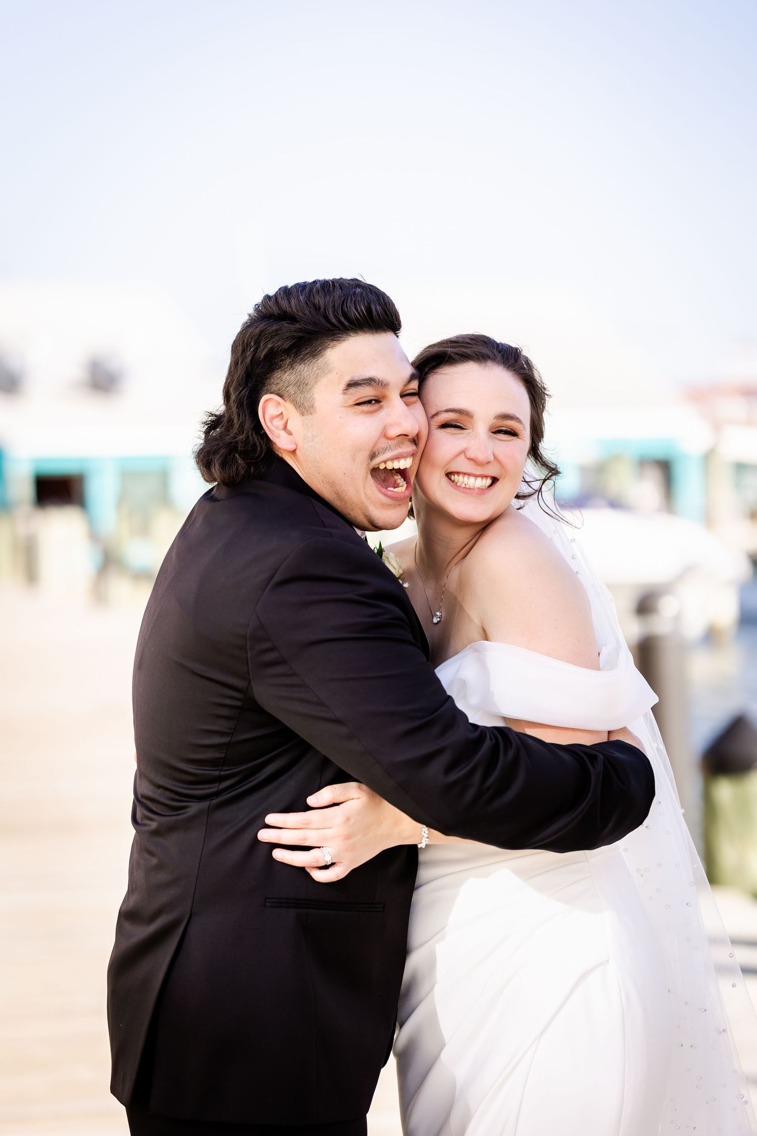 Two happy people in formal wedding attire laugh and embrace joyfully during an outdoor celebration.