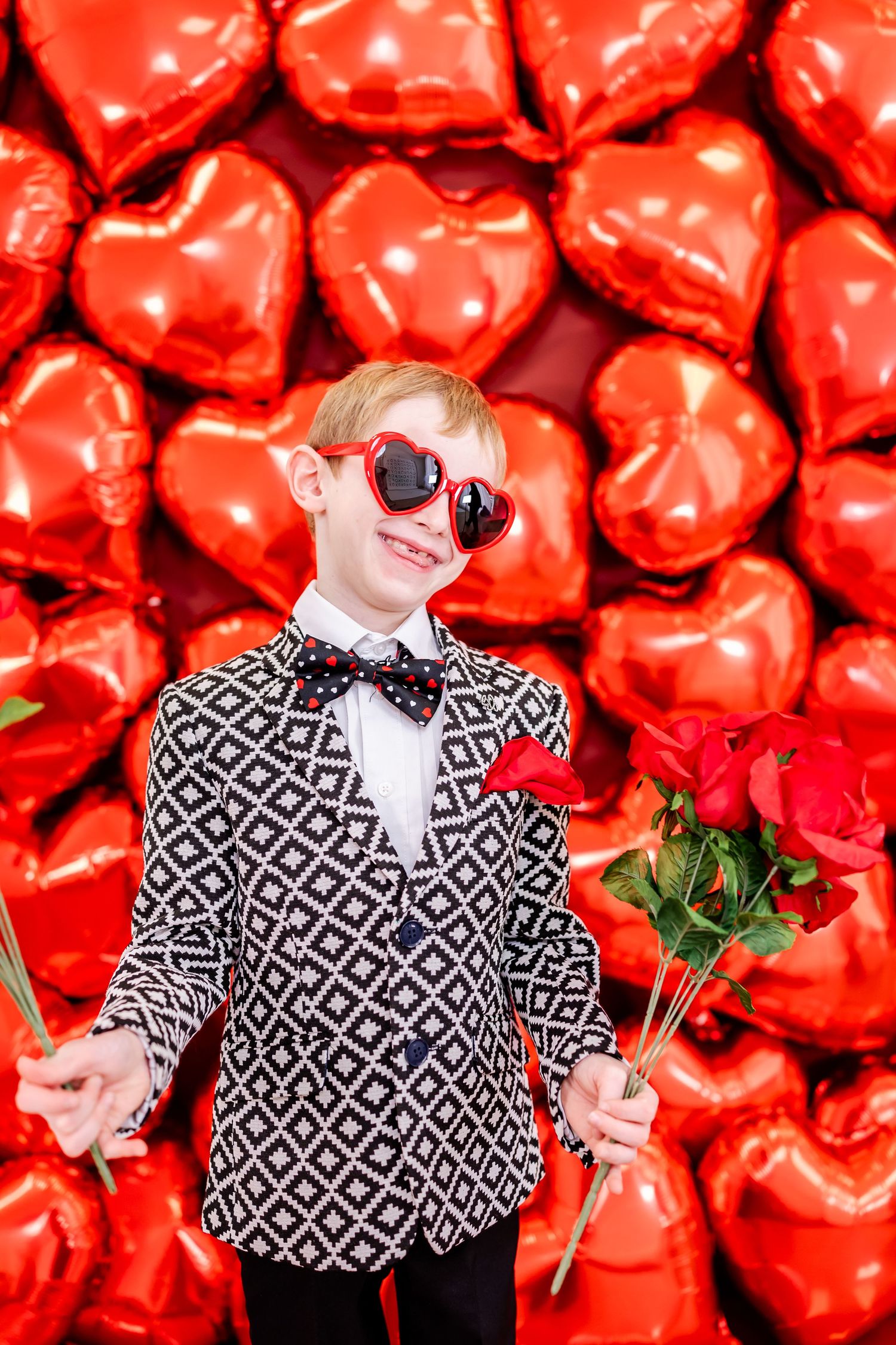 Stylish figure in geometric print blazer and heart sunglasses poses with roses against red heart balloon background.