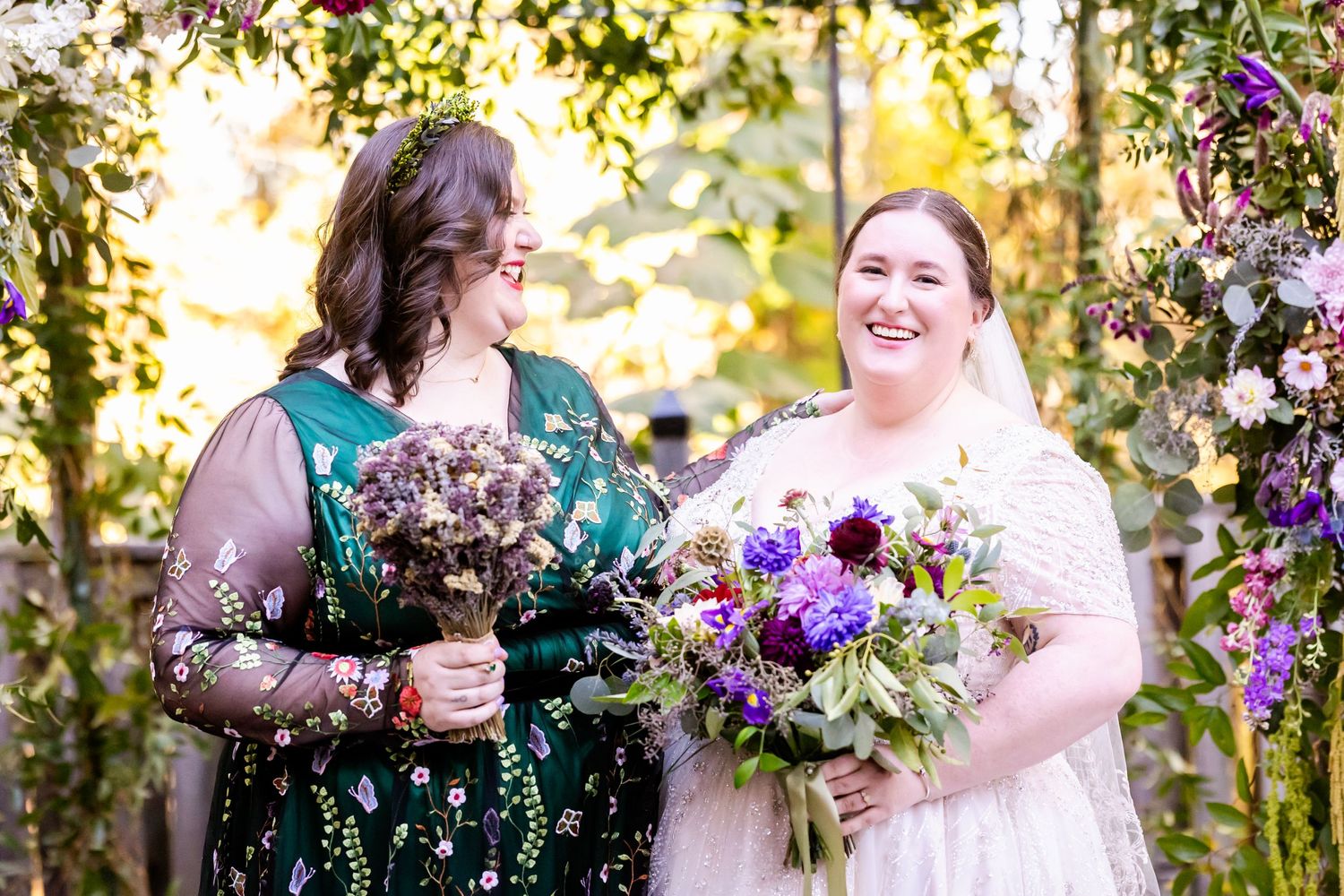 Two wedding attendees share a joyful moment surrounded by purple floral arrangements.