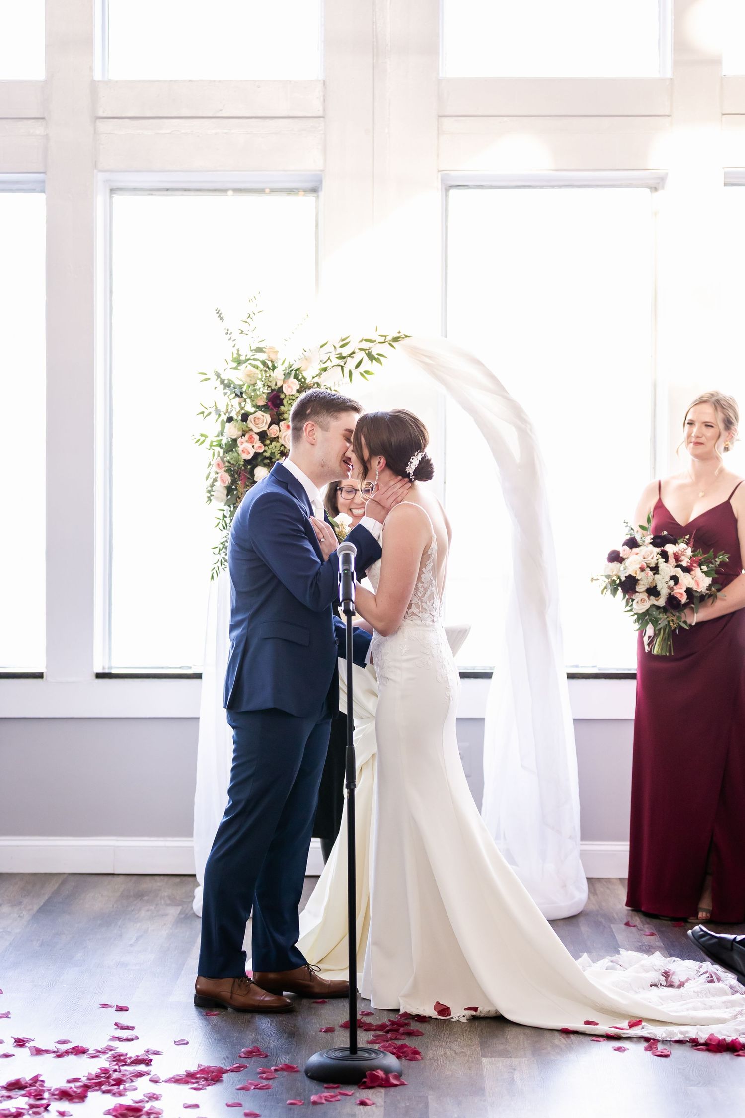 Wedding ceremony under large windows with floral arch and scattered rose petals.
