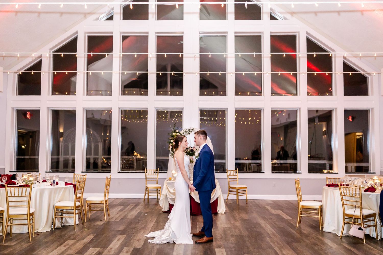 Large bright wedding venue with white walls, windows and wooden floors set up for a reception.