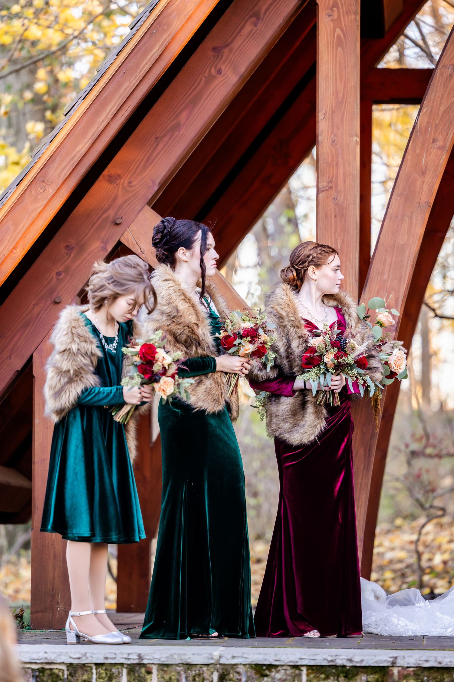 Bridesmaids in emerald green and burgundy velvet dresses pose playfully in a rustic wooden gazebo during autumn.