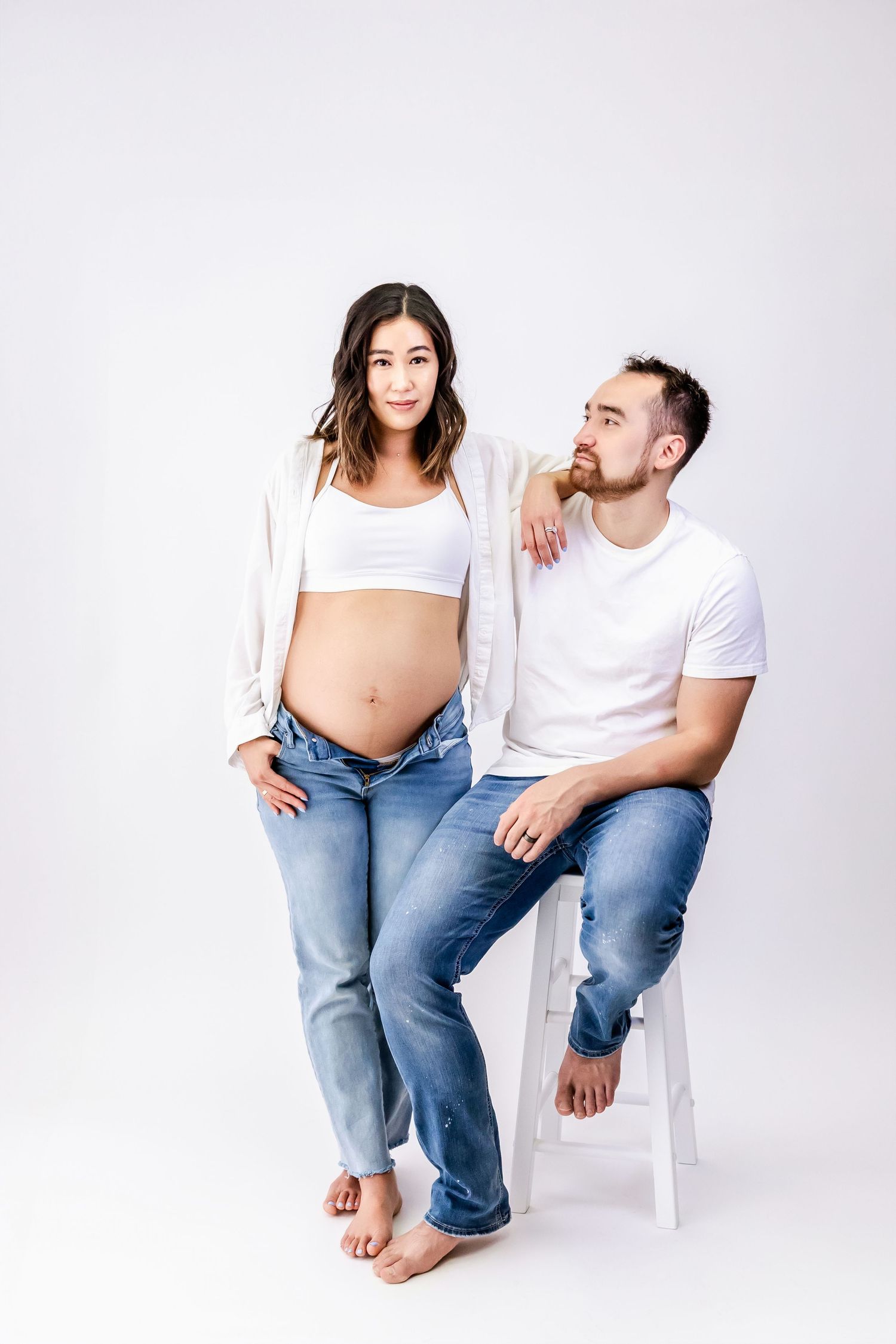 A couple in jeans and white tops sit casually on white chairs against a light background.